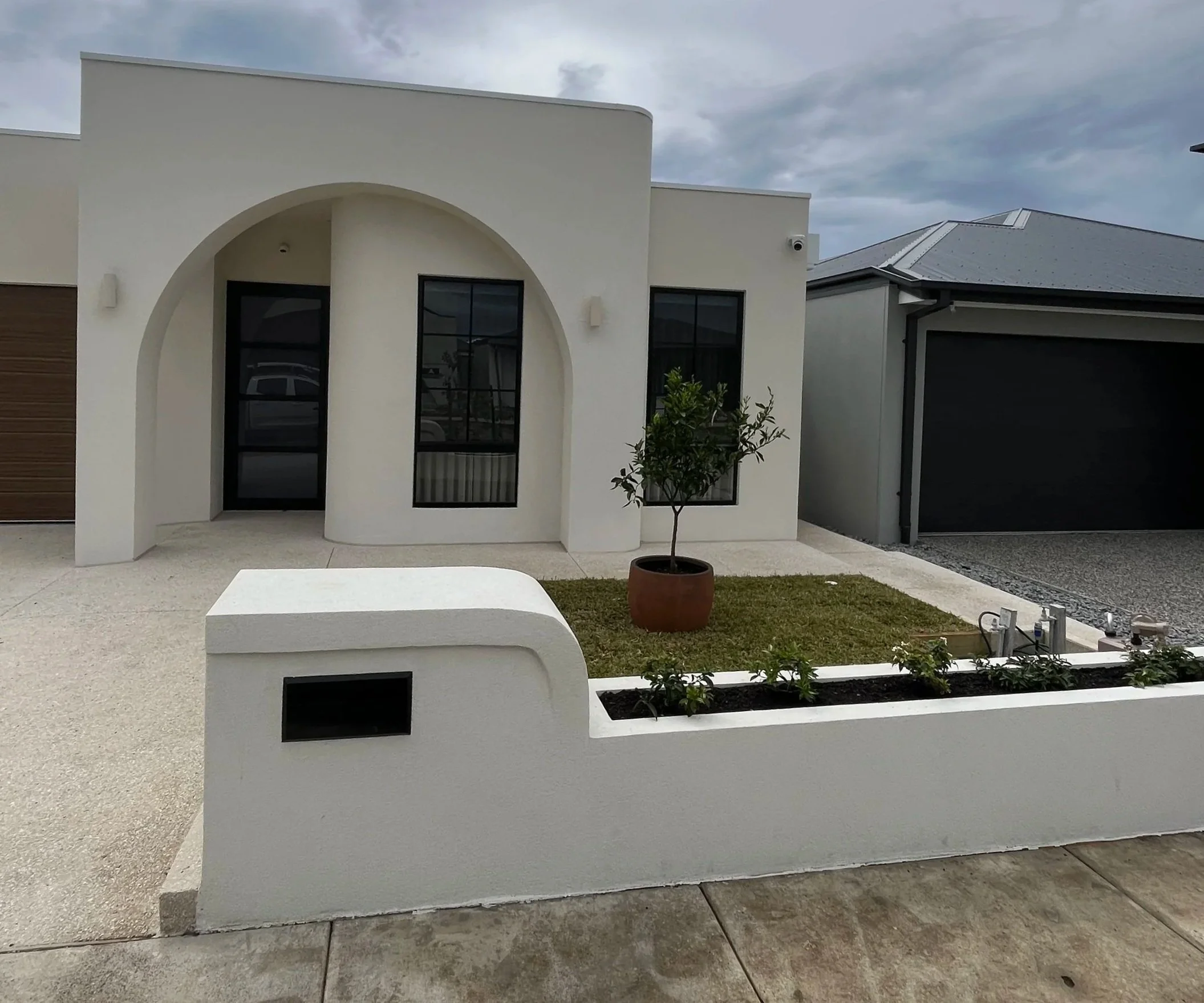 Modern white house with arched entryway, large black windows, a small tree in a pot, and a white concrete bench outside. Overcast sky in background.