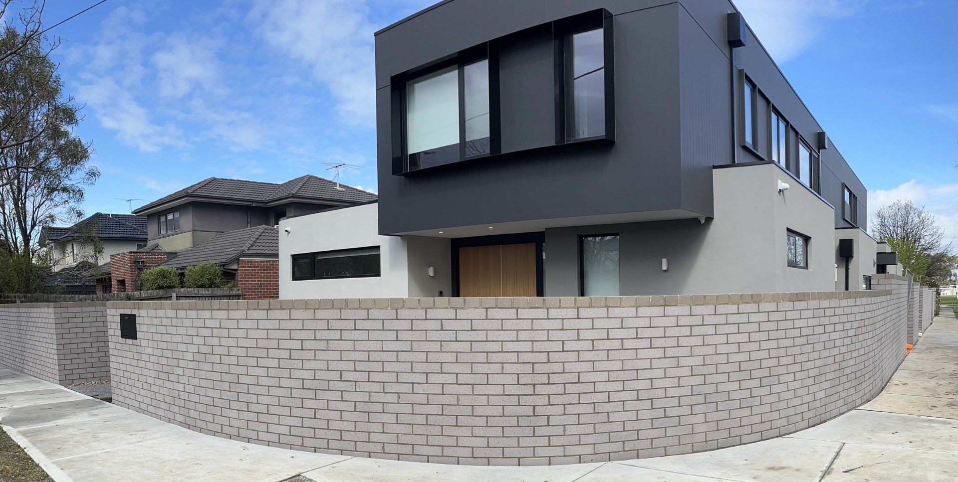 Modern, two-story house with gray and white exterior, large windows, and a brick perimeter wall, in a suburban neighborhood.