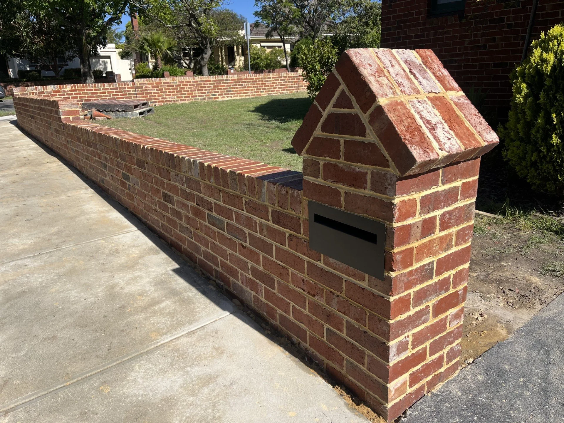 A brick mailbox on a concrete sidewalk with a brick wall surrounding a lawn, trees, and houses in the background.