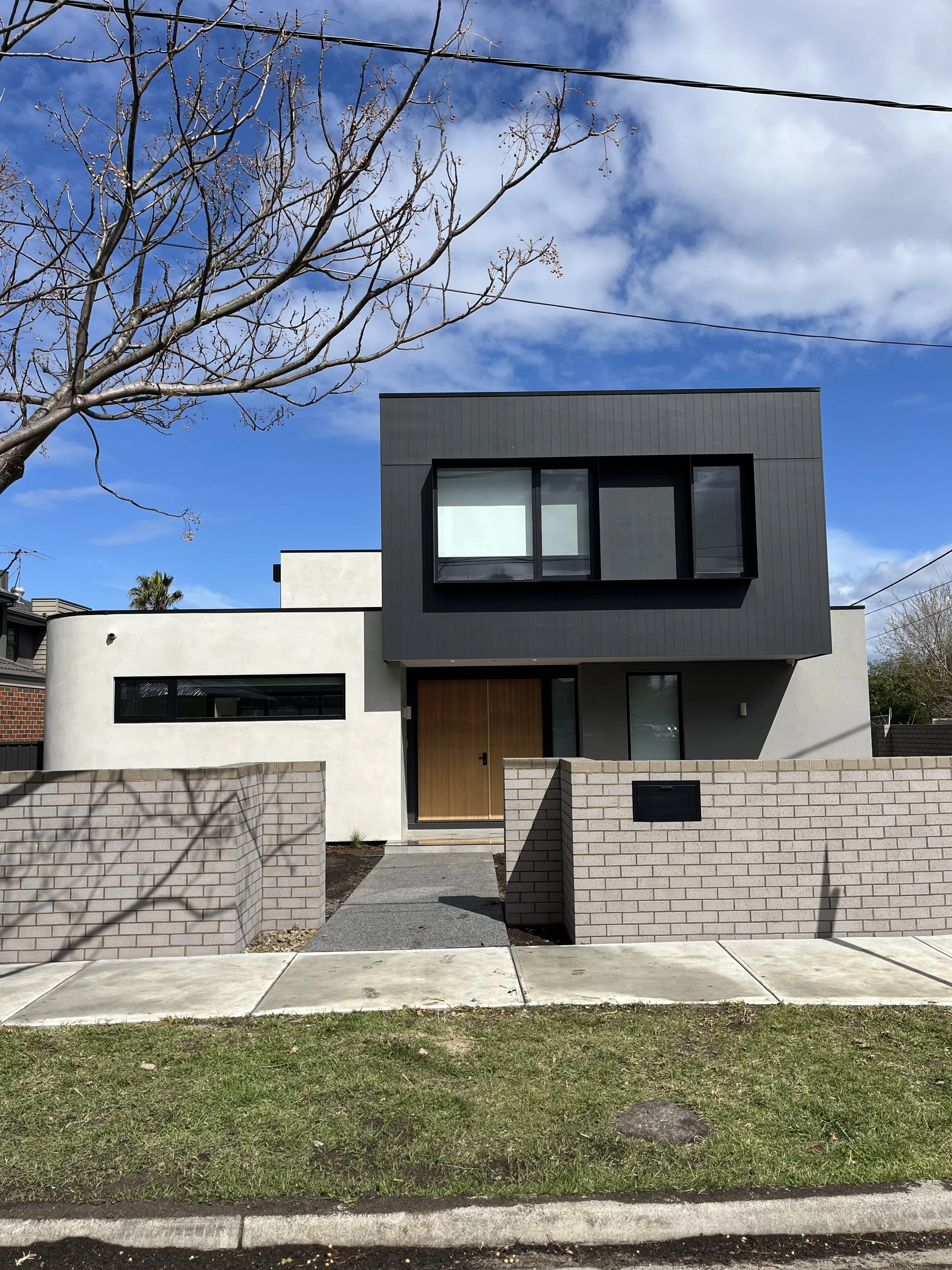 Modern two-story house with white and dark gray exterior, large windows, a wooden front door, and a low brick wall in front. A leafless tree is in the foreground, with a blue sky and a few clouds above.