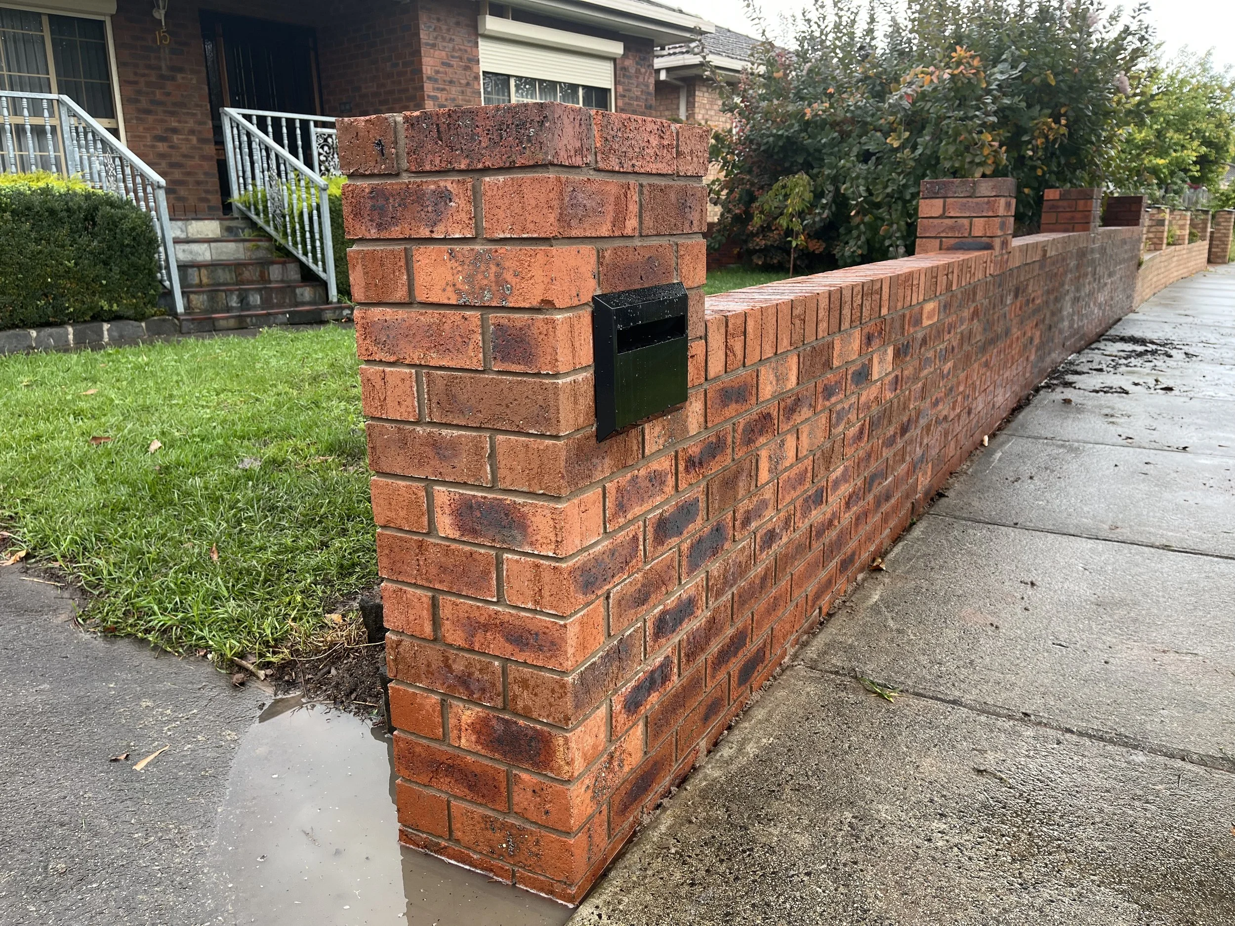 Red brick wall with a small black mailbox, adjacent to a sidewalk and grass, in a residential neighborhood with houses and trees.