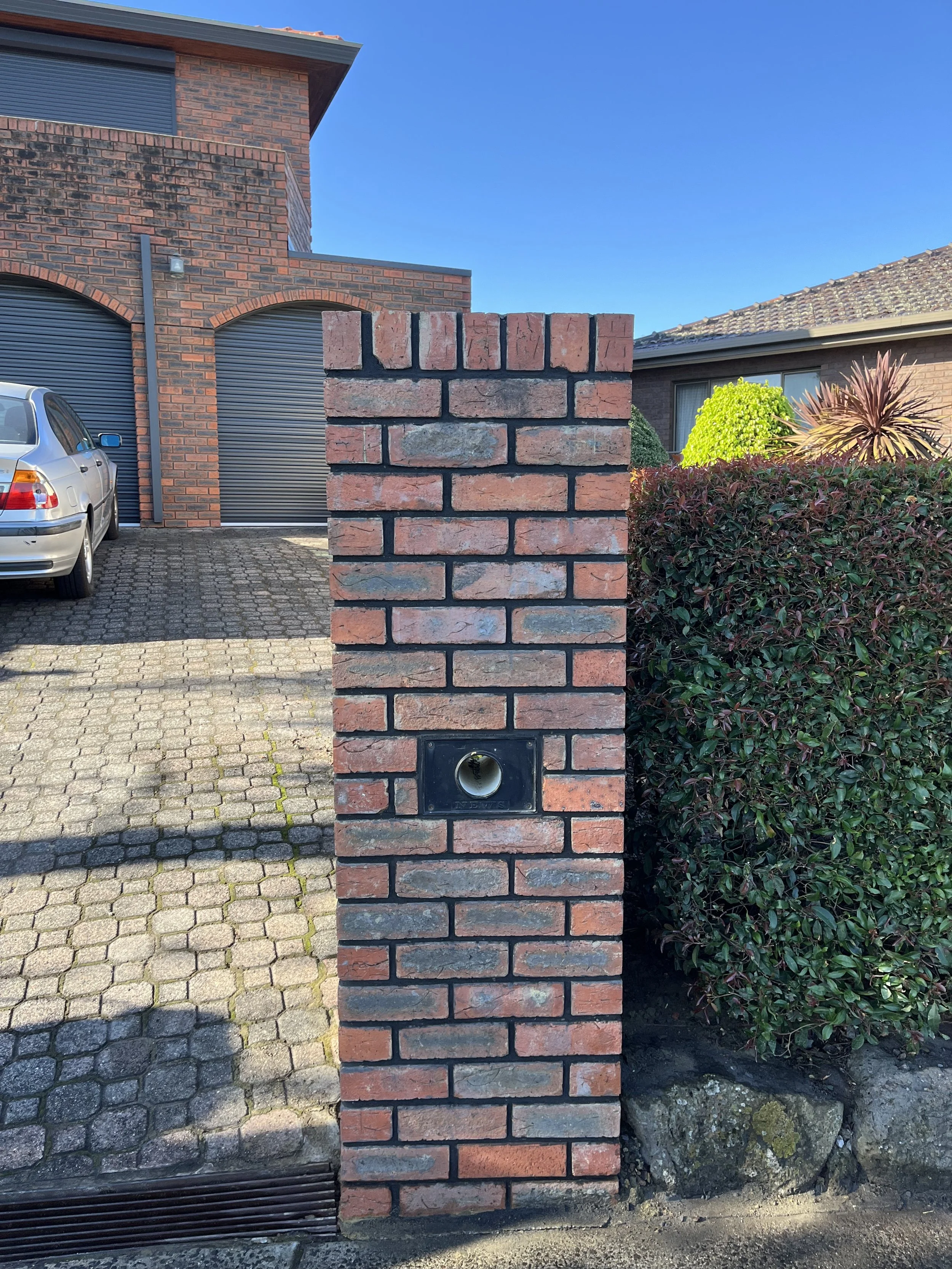 A brick mailbox with a metal mailbox slot, situated in front of a residential house. The house has brick walls, a garage with a metal door, and a car parked in the driveway. There are bushes and plants beside the mailbox.