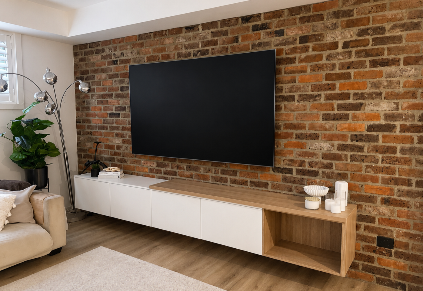 Living room with brick accent wall, mounted flat-screen TV, and modern white and wood entertainment console with decorative candles, a bowl, books, a plant, and a lamp nearby.