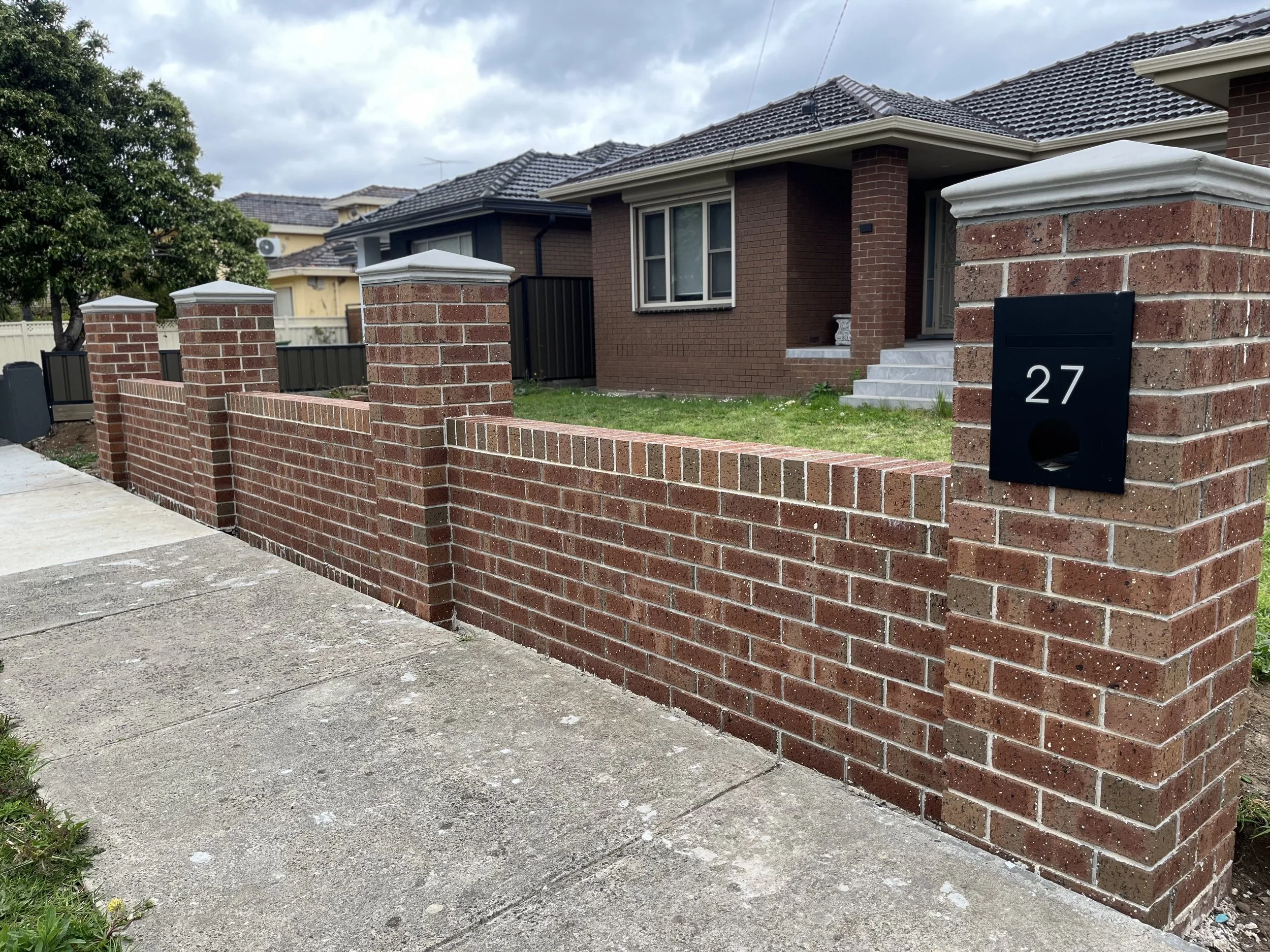 Front view of a brick house with a brick wall fence and white mailbox with number 27 in black, gray steps leading to the front door, cloudy sky, lush green grass, and trees in the background.