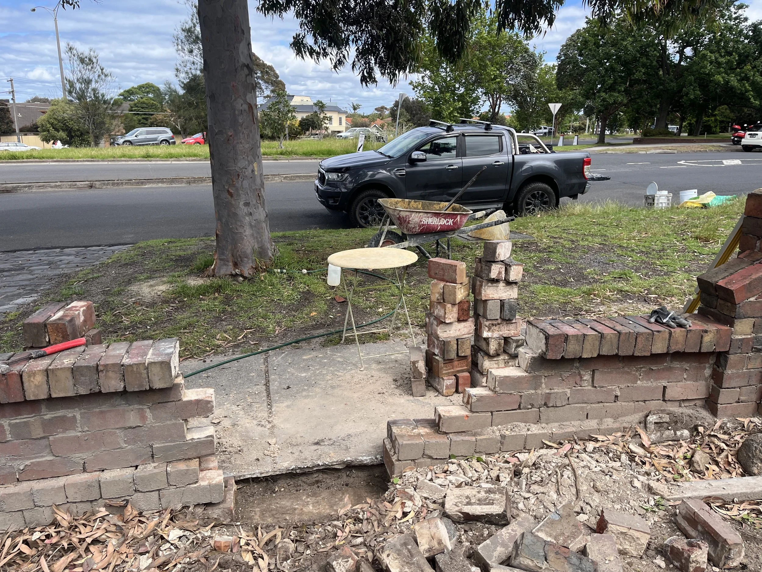 A construction site with a partially built brick wall, construction materials, and a black pickup truck parked on the street. Background shows trees, cars, and houses under a partly cloudy sky.