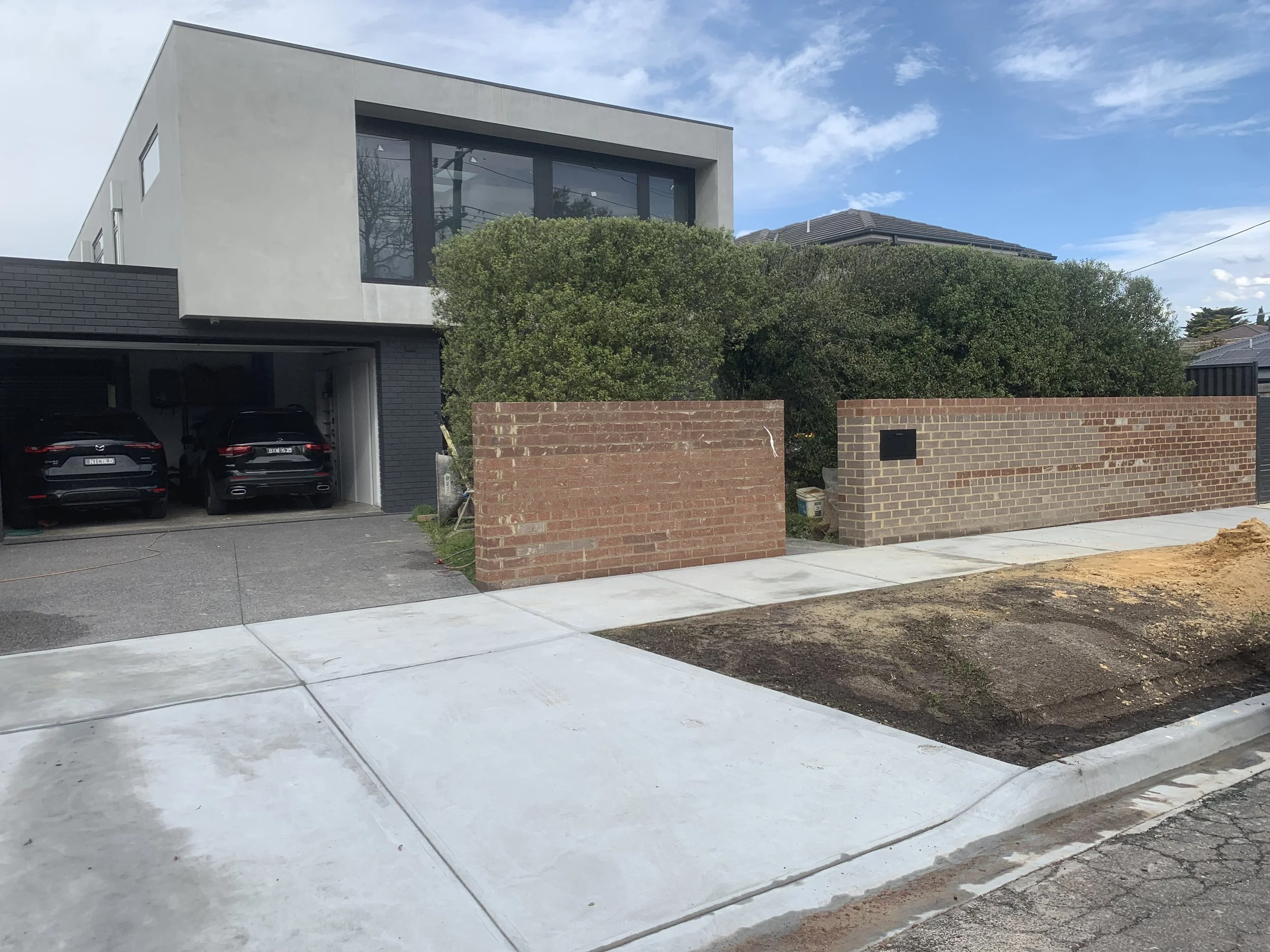 Modern two-story house with large windows, a brick wall, and a driveway with two black cars parked inside a garage. Landscaping includes green bushes and a partly cloudy sky.