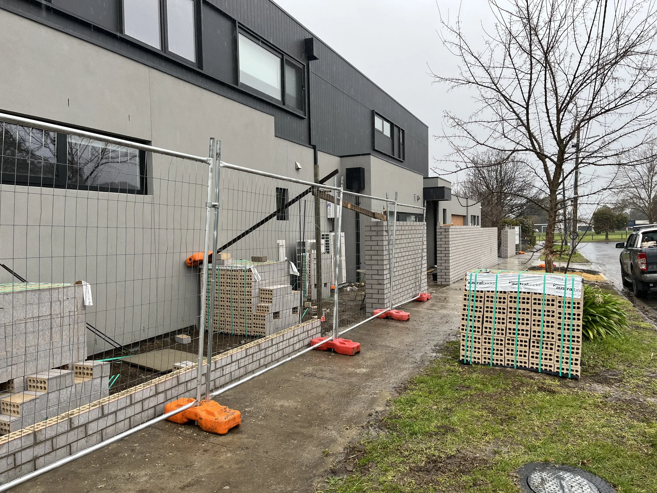 Construction site in front of a modern building with black and beige exterior walls, with gray overcast sky, trees, parked cars, and building materials and fencing around the area.