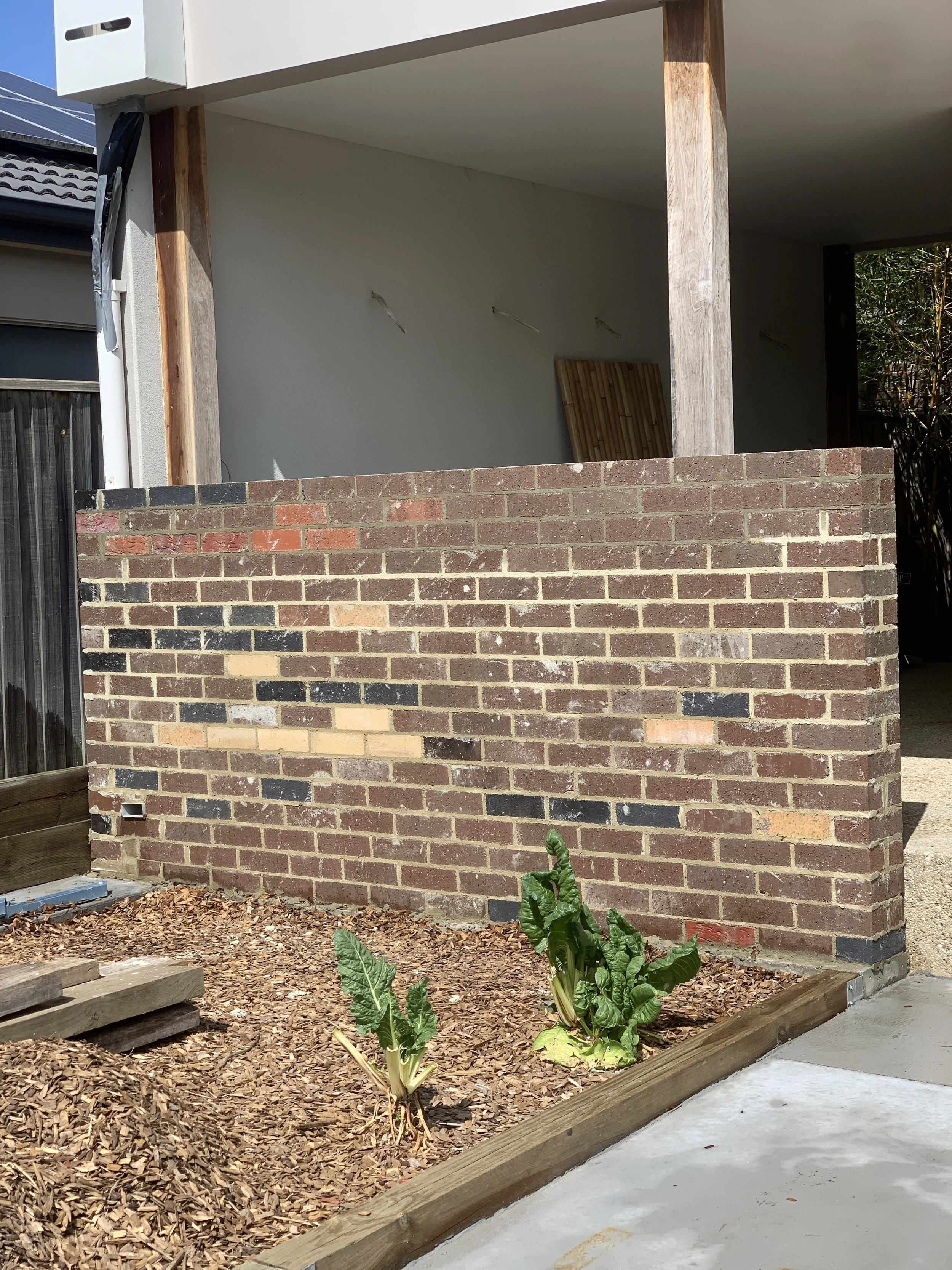 A small unfinished outdoor patio with a brick wall, wooden posts, and a white ceiling. There are some plants and mulch in the garden bed, with wooden edging and a concrete path in the foreground. Part of a house with solar panels on the roof and a sl