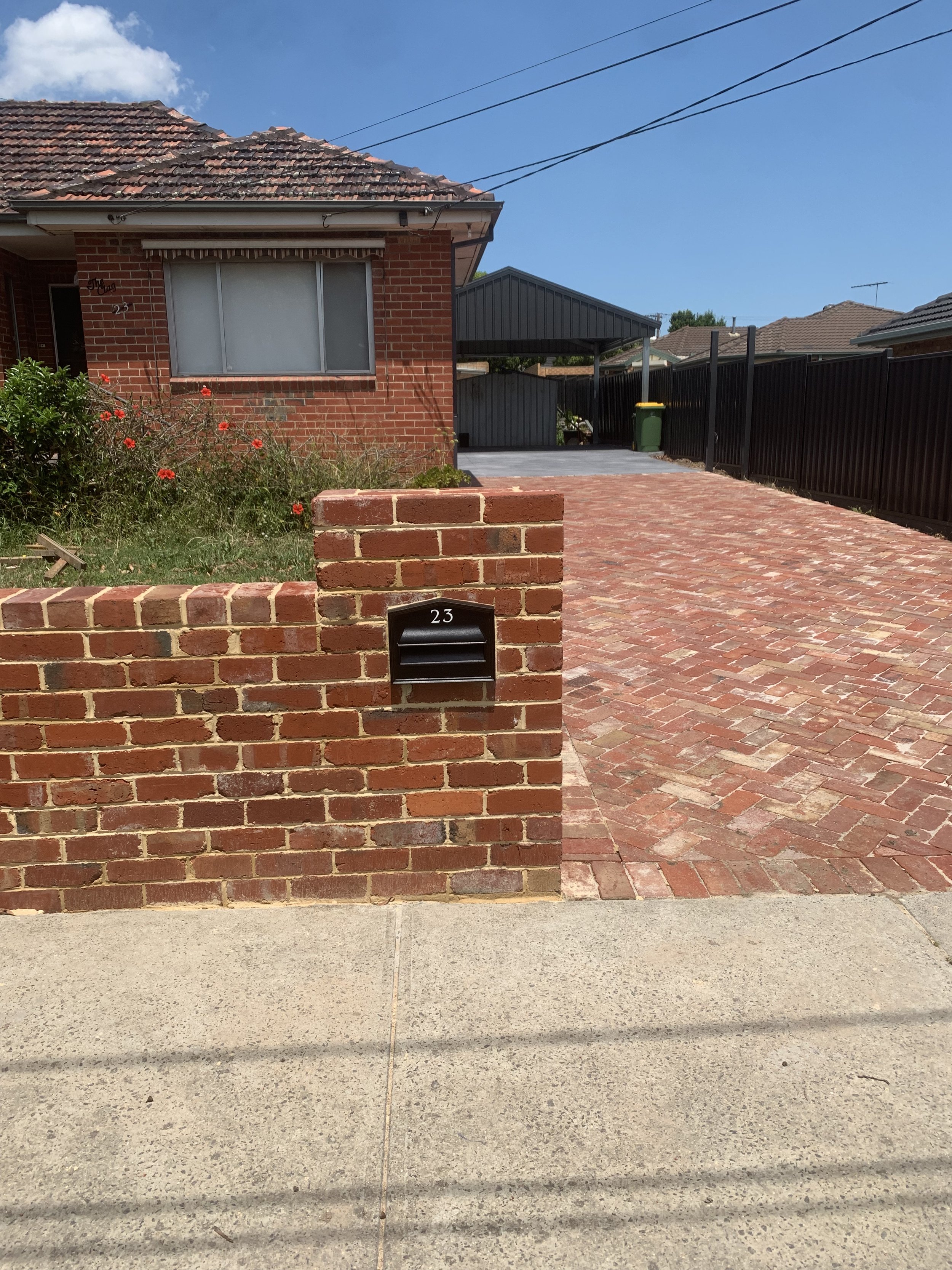 A house with a brick facade, a driveway with brick pavers, a small garden with red flowers, a brick mailbox with the number 23, and a black fence. The sky is blue with some clouds.