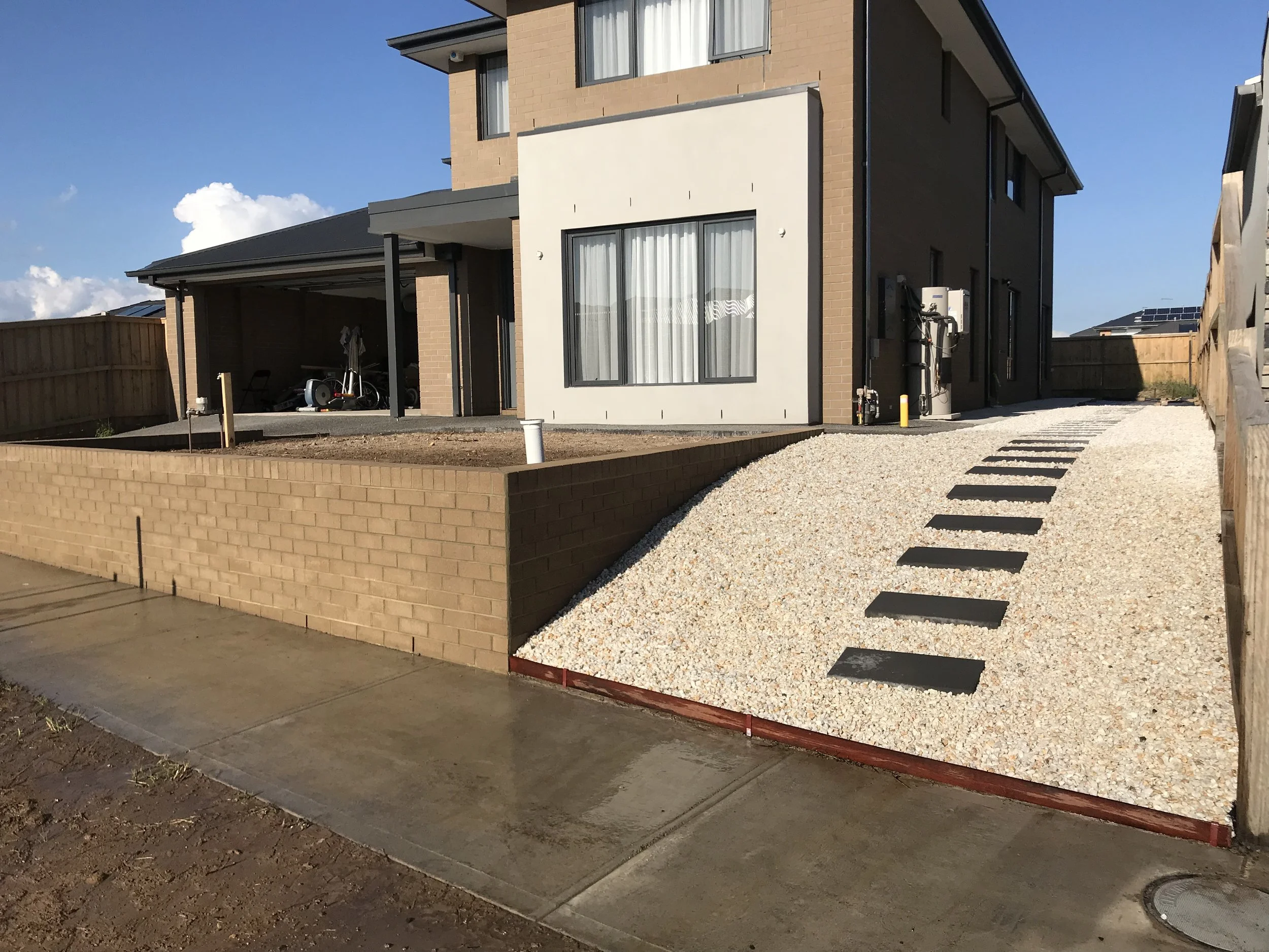 A modern, multi-story residential building with exterior brick and painted walls, large windows with curtains, a carport with a bicycle and other items stored underneath, and a gravel pathway with black stepping stones leading to the entrance. A brick boundary wall and a fence surround the property with neighboring houses visible in the background, under a blue sky with some clouds.