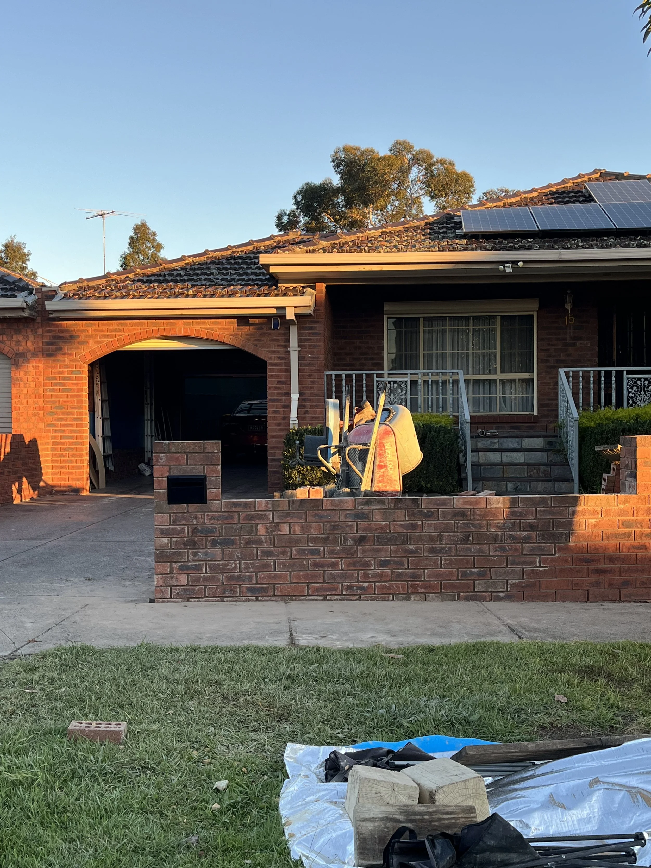 A brick house with solar panels on the roof, a garage, a small stairs leading to the front door, a lawn with some grass, and construction materials in the foreground at sunset.
