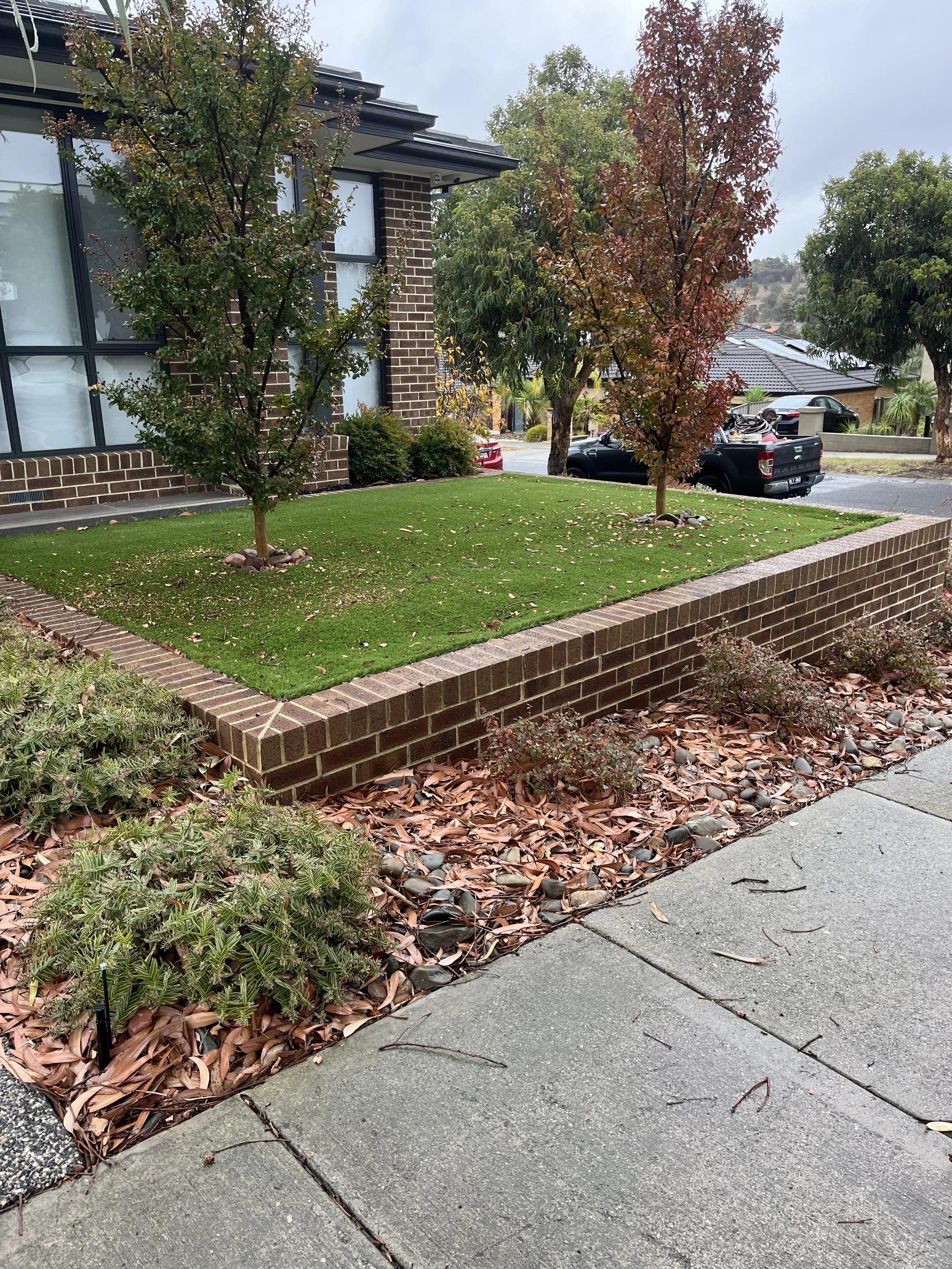 A well-maintained front yard with a small brick wall border, mulch with fallen leaves, and a green lawn with two young trees, on a cloudy day in a suburban neighborhood.