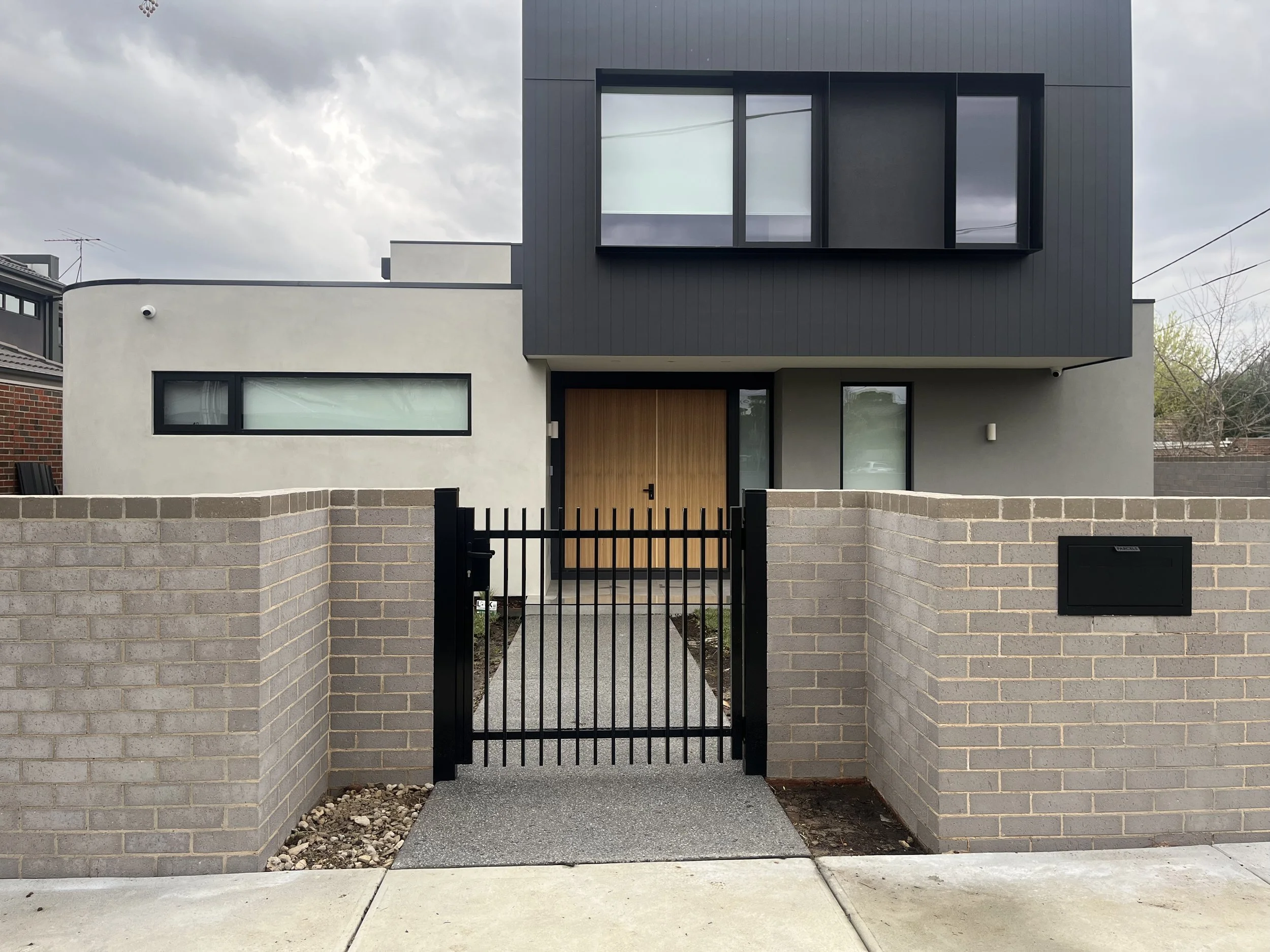 Modern house with a gray upper section, white lower section, large windows, and a wooden front door, enclosed by a brick fence and black gate, under a cloudy sky.