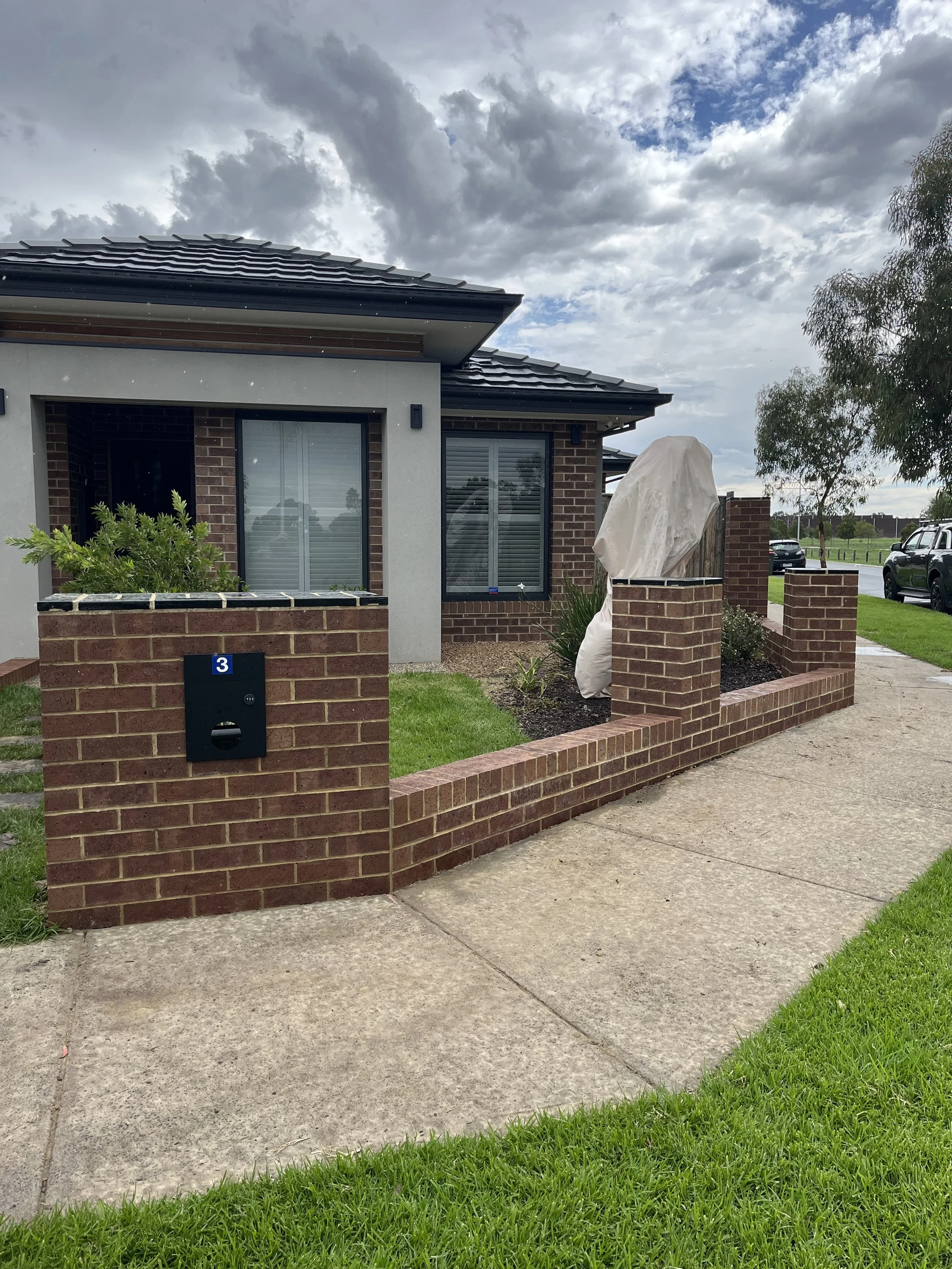 Front view of a brick house with a porch, a brick wall with a mailbox, and a small landscaped garden. The sky is cloudy, with patches of blue sky visible.