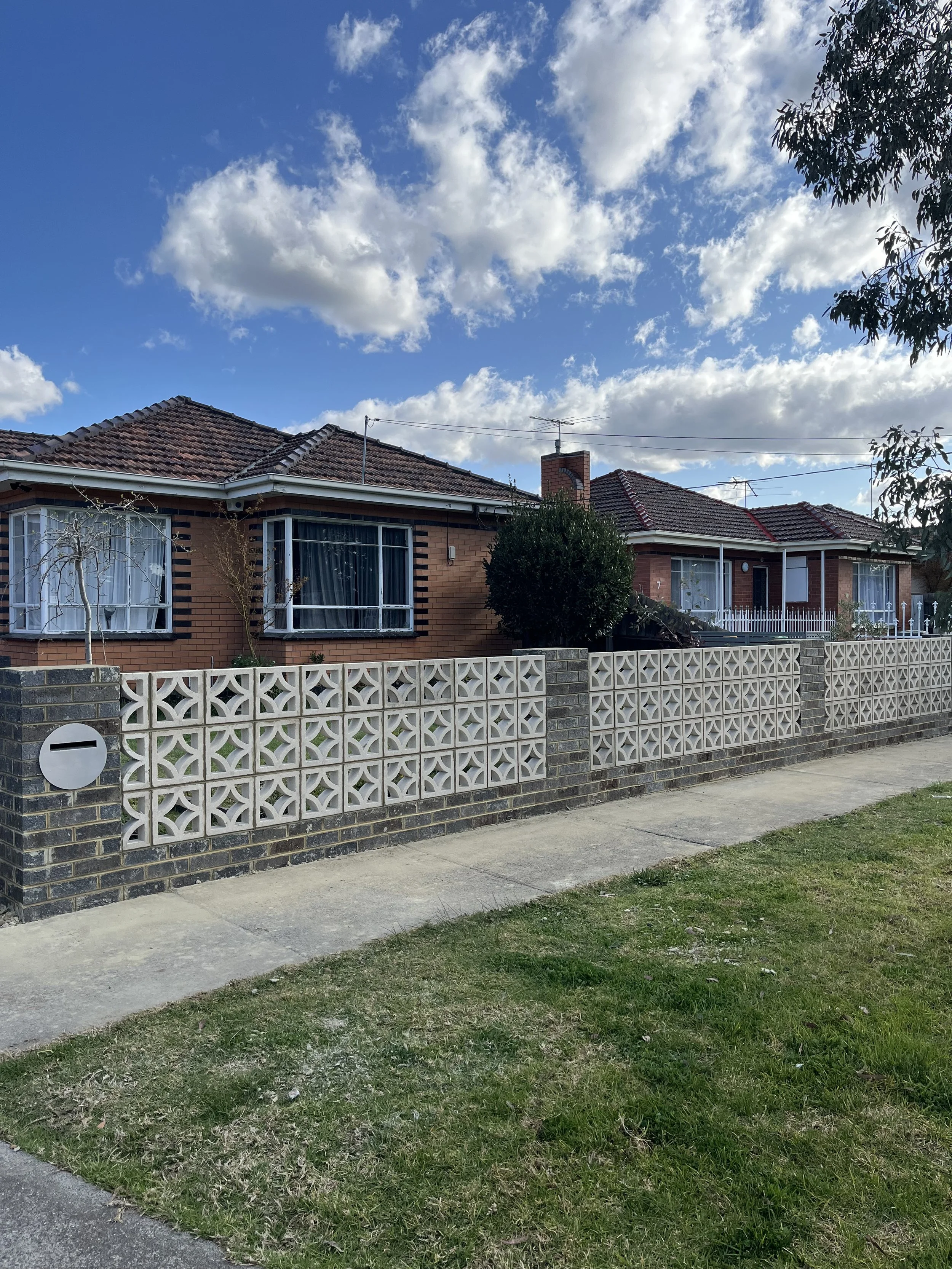 A suburban house with a red brick exterior and a tiled roof, surrounded by a decorative concrete fence and a grassy yard with trees.