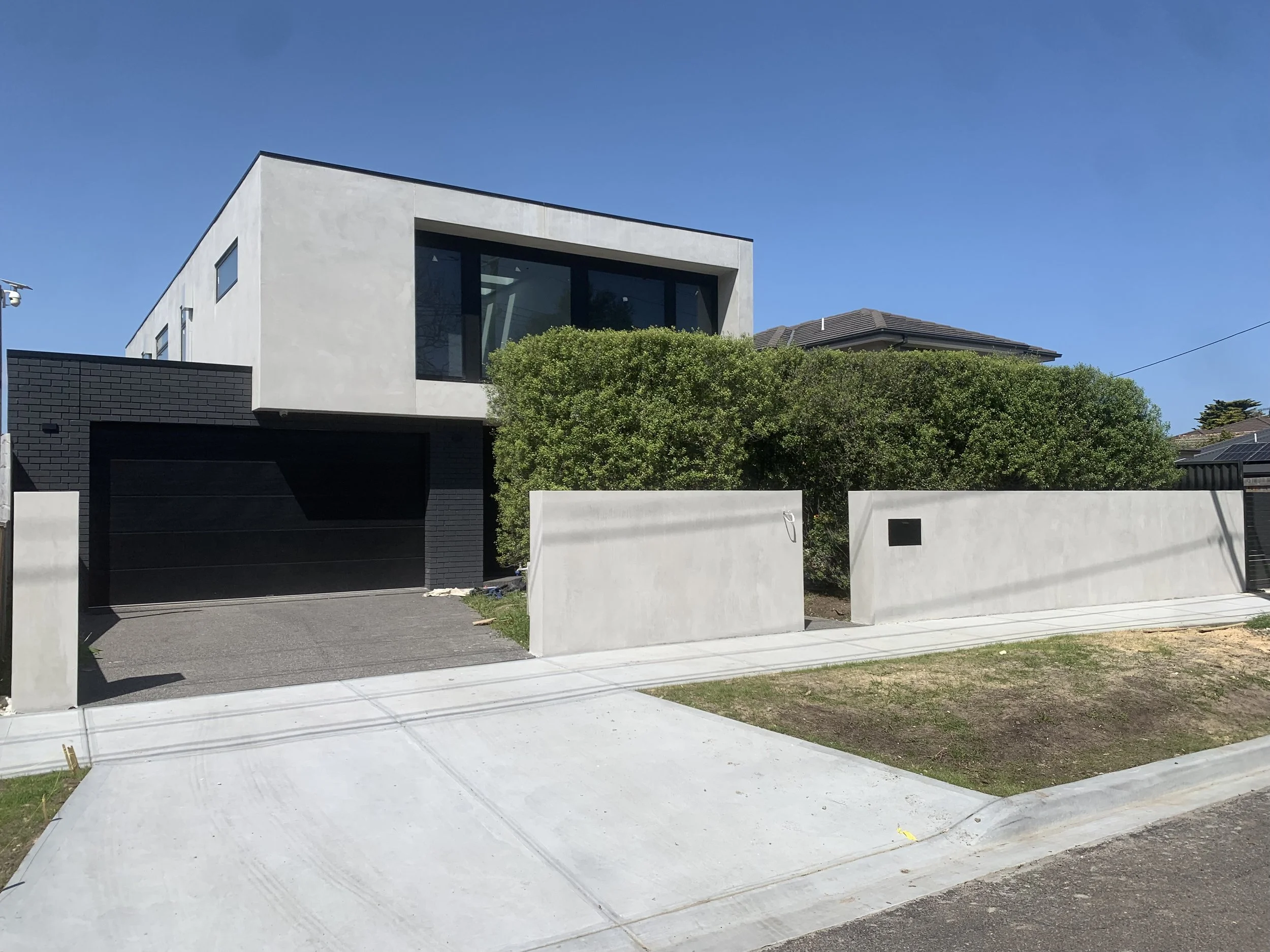 Modern two-story house with white exterior walls, large dark windows, a black garage door, and surrounded by a white wall and green bushes. Clear blue sky in the background.