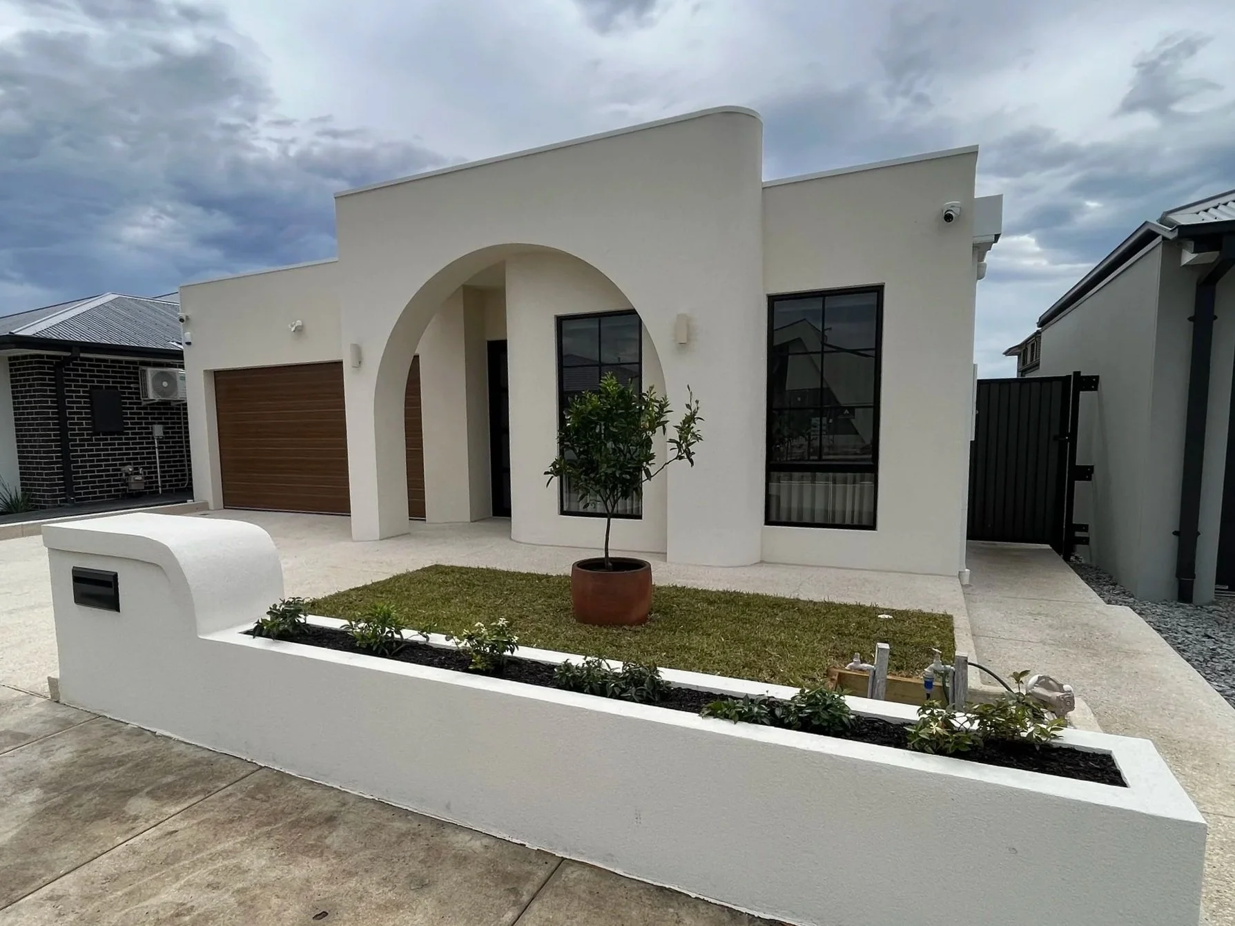 Modern white house with curved architectural features, large windows, front door, small tree in a pot, driveway, and a garden with a white border wall and small plants.