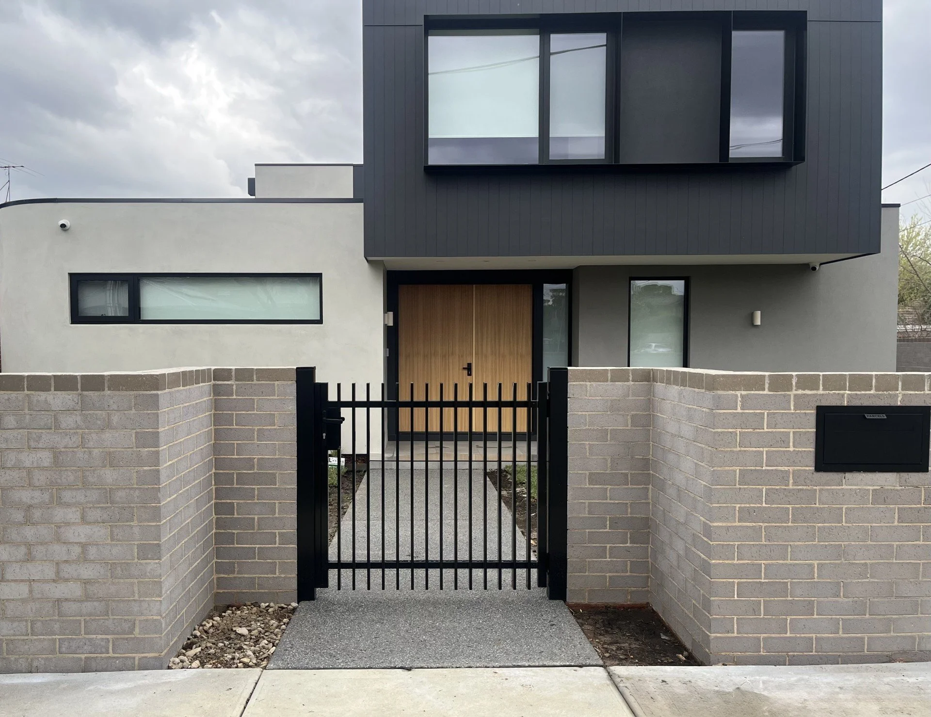 Modern house with gray and white exterior, large windows, wooden front door, brick wall, black gate, and mailbox outside, under cloudy sky.