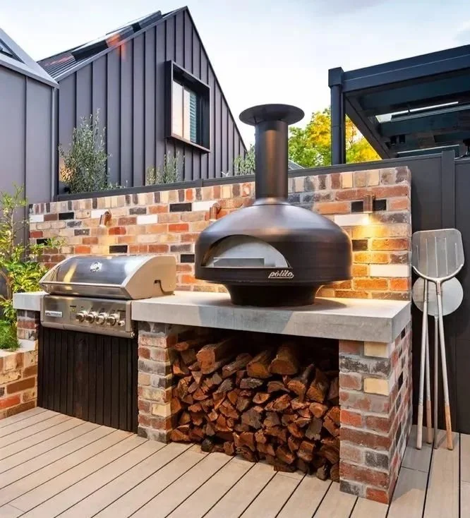 An outdoor kitchen with a brick and wood fire oven, a BBQ grill, a wood storage area, and folding chairs. There are plants and a modern building in the background.