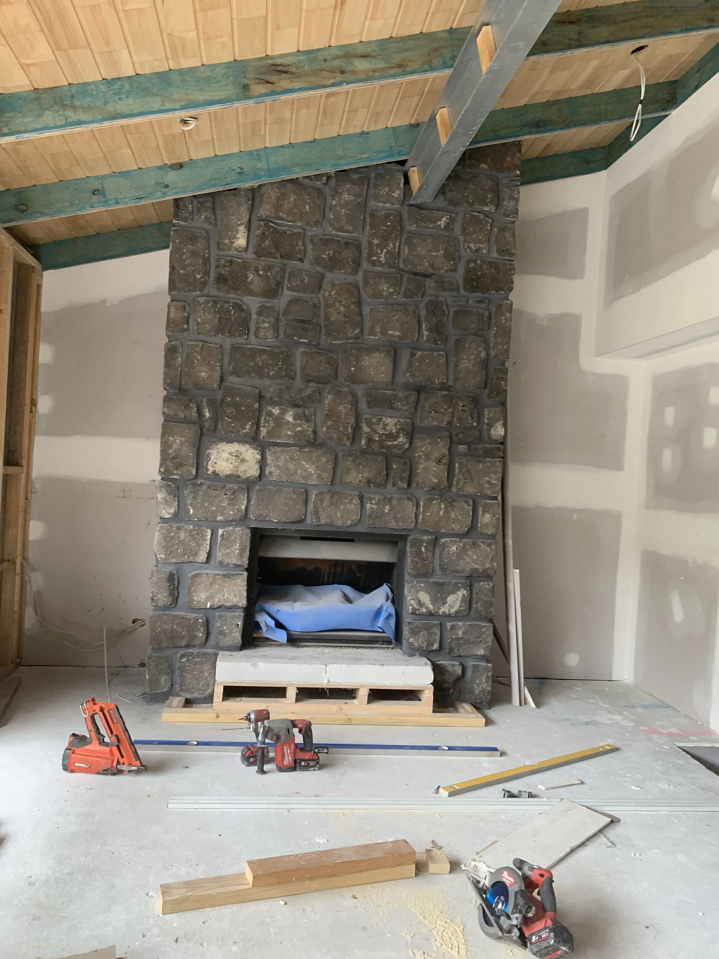 Inside view of a room under construction with a stone fireplace surround, construction tools like drills, saws, and a level on the floor, and unfinished walls and ceiling with exposed framing and insulation.
