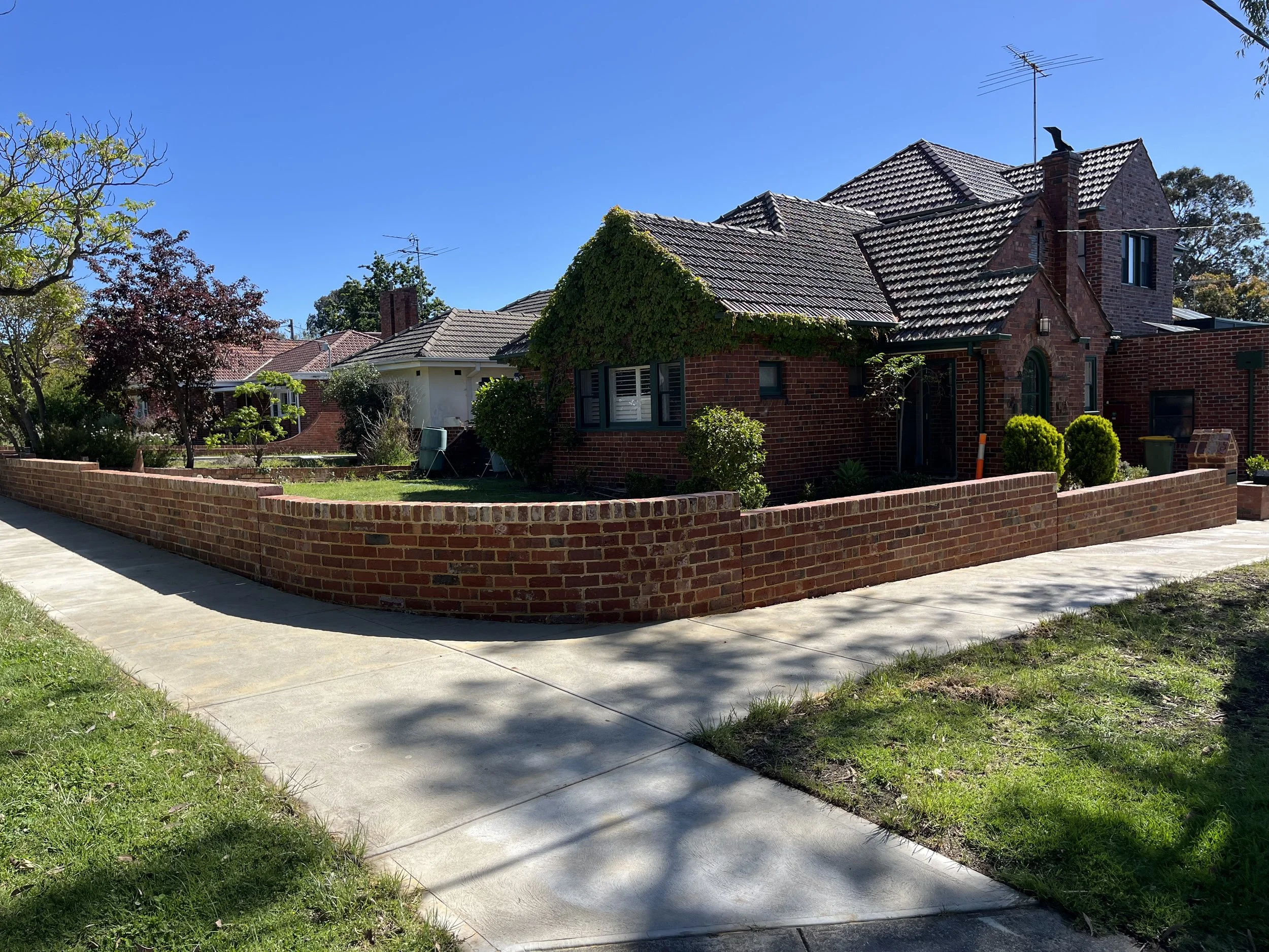 A residential neighborhood with brick houses, some with ivy-covered walls, surrounded by trees and a brick fence, under a clear blue sky.