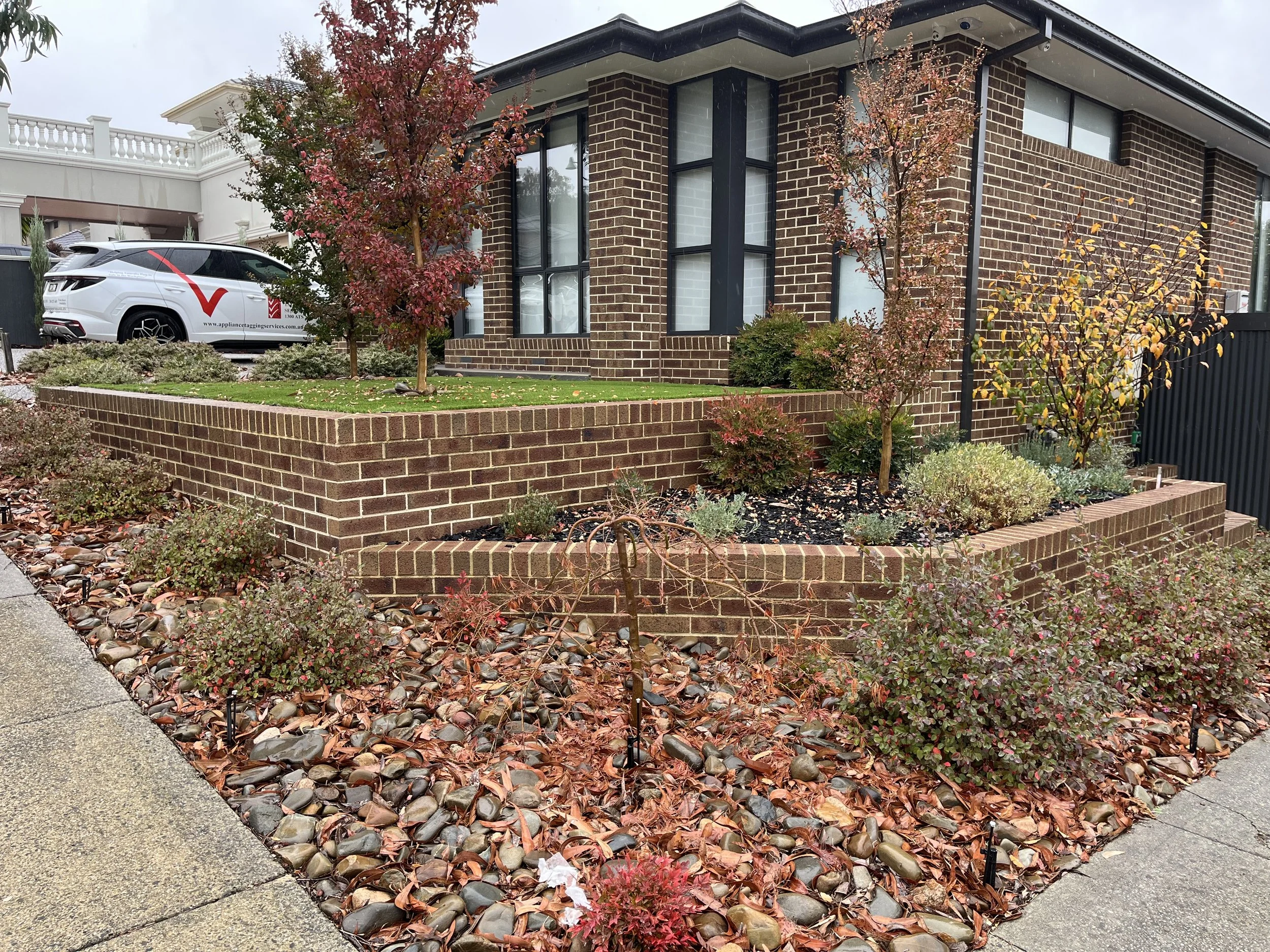 A brick residential building with large windows, surrounded by landscaped gardens with small trees, bushes, and mulch, on a cloudy day.