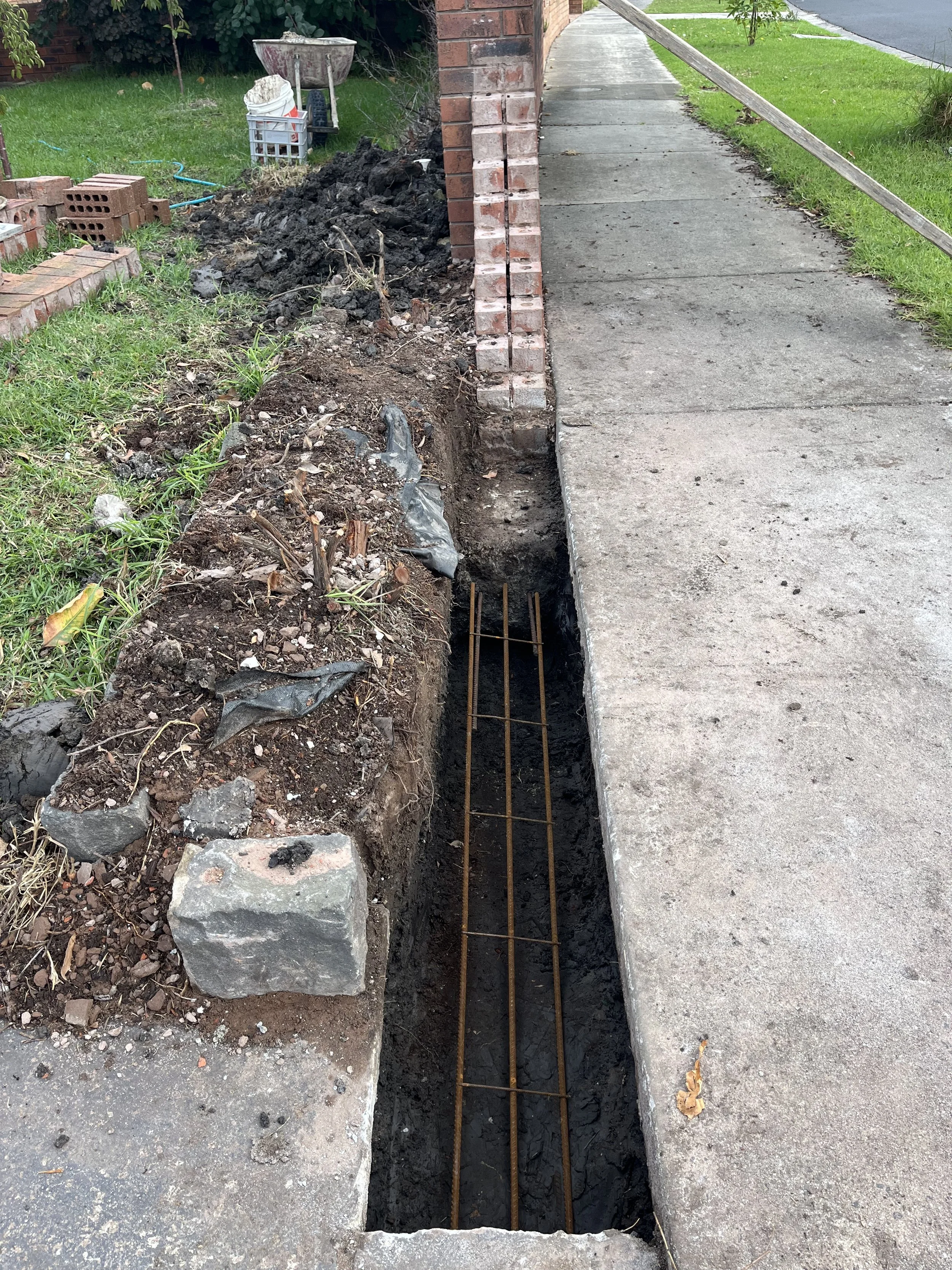 Sidewalk under construction with a trench dug next to it, exposing rebar. There's a brick wall on the left, green grass, and sidewalk on the right. Construction materials and tools are visible.