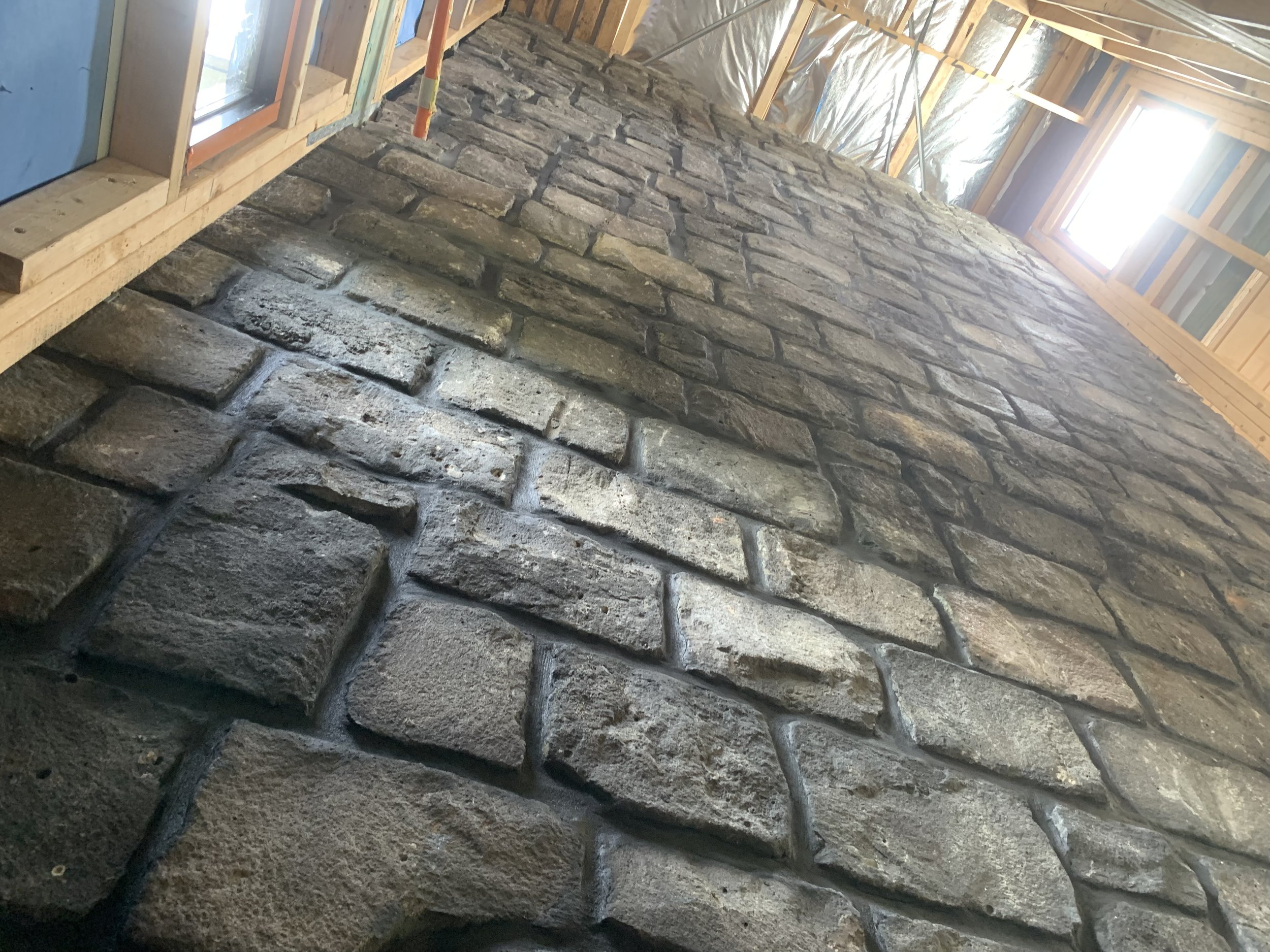 Interior view of a building under construction with a partially built stone wall and exposed wooden framing, insulation, and two windows allowing natural light.