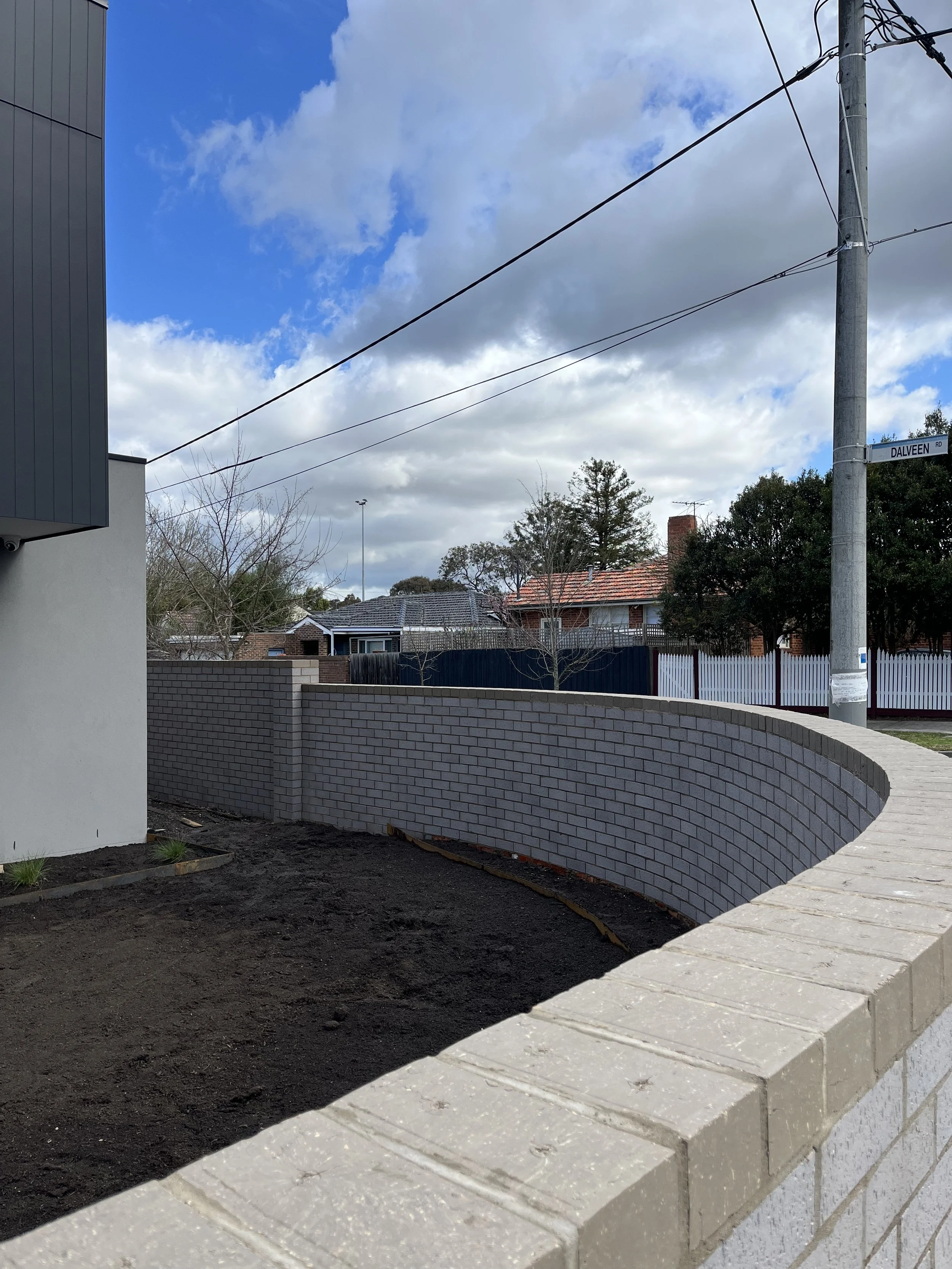A curved brick wall in front of a house with a white fence, some trees, and a partly cloudy sky.