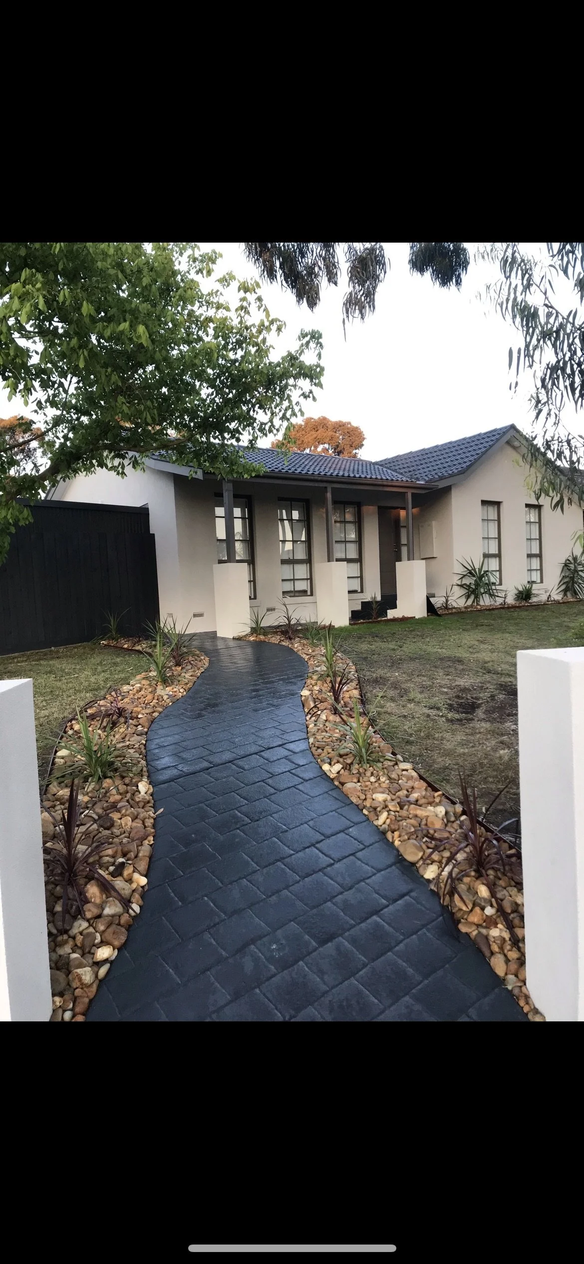 A modern house with a curved black paved walkway leading to the front entrance, bordered by small rocks and plants, surrounded by trees and greenery.
