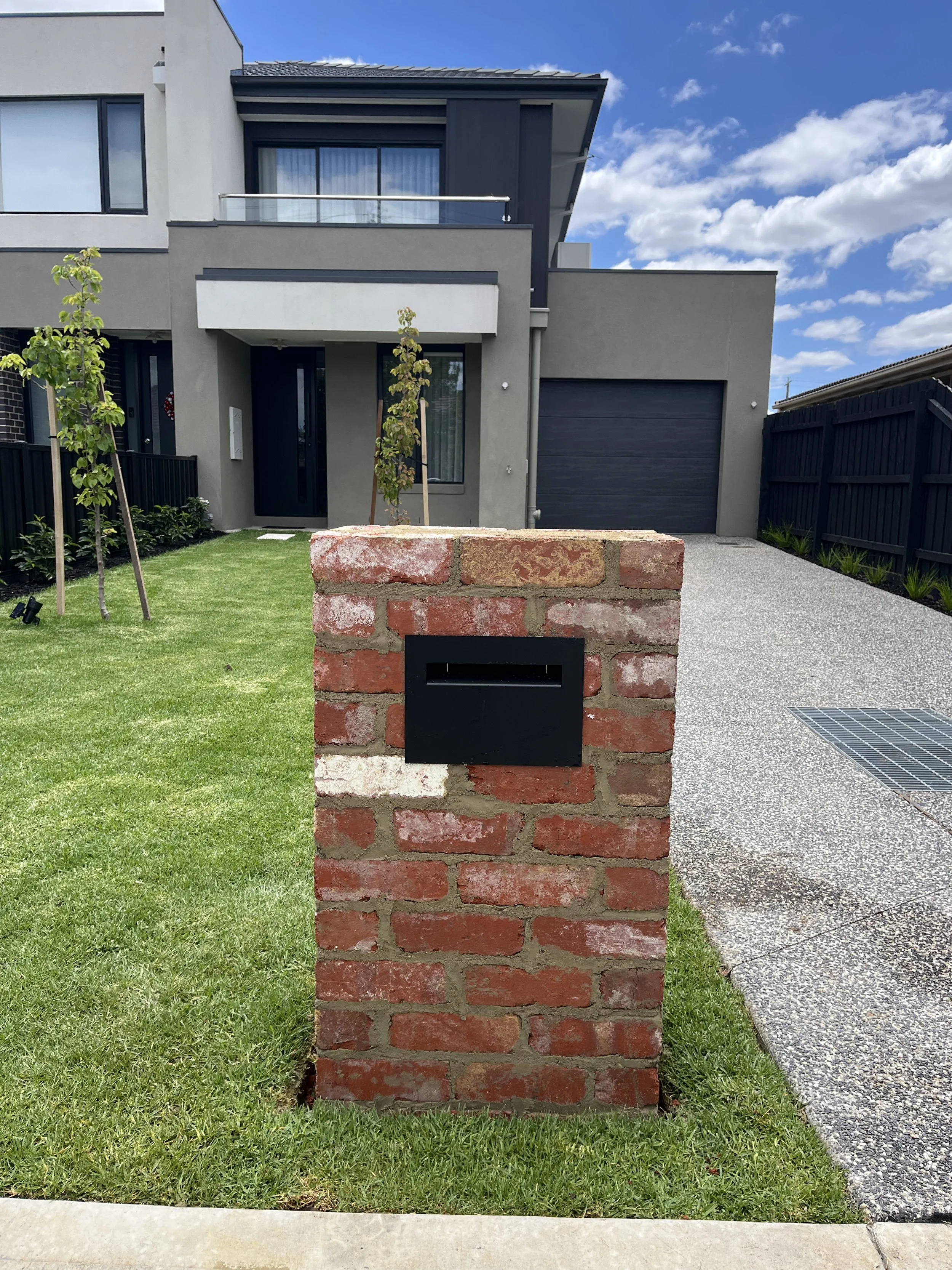Modern house with gray exterior, black garage door, small front yard with green grass, young trees, brick mailbox, and a bright blue sky with scattered clouds.