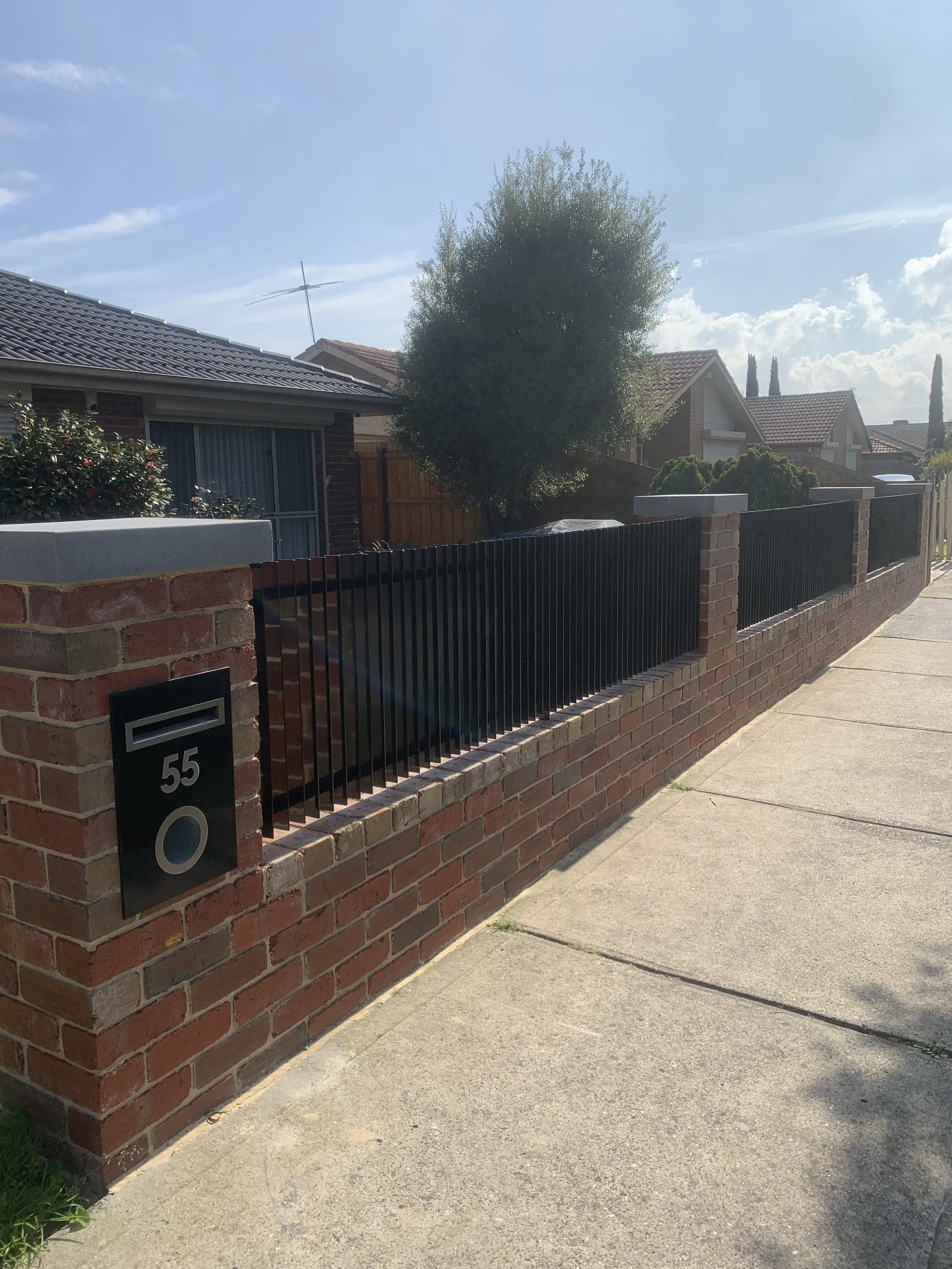 Residential front yard with brick fence, black metal fence, trees, and single-story houses under a blue sky with some clouds.