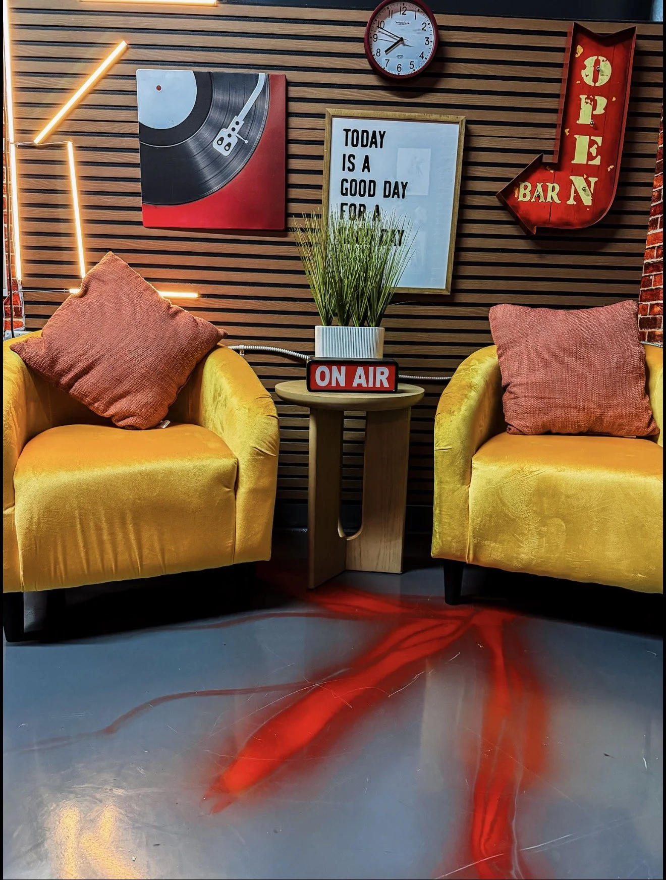 A cozy bar setting with two yellow armchairs with pink cushions, a small round wooden table between them, a potted plant, and various wall decorations including a clock, a sign that reads 'Today is a good day for a drink', a vinyl record art piece, and a red 'Open Bar' arrow sign. The floor has a red abstract pattern.