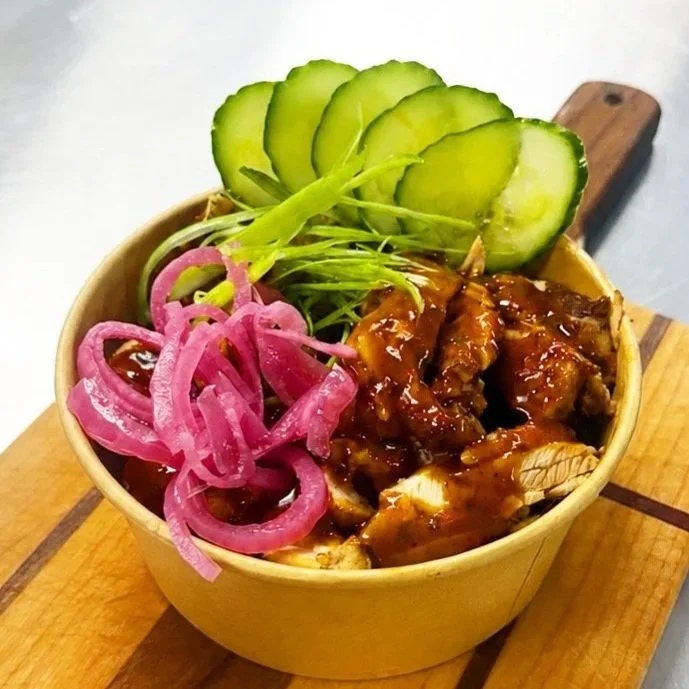 Bowl of barbecue chicken with sliced cucumber, pickled pink onions, and green garnishes on a wooden board.
