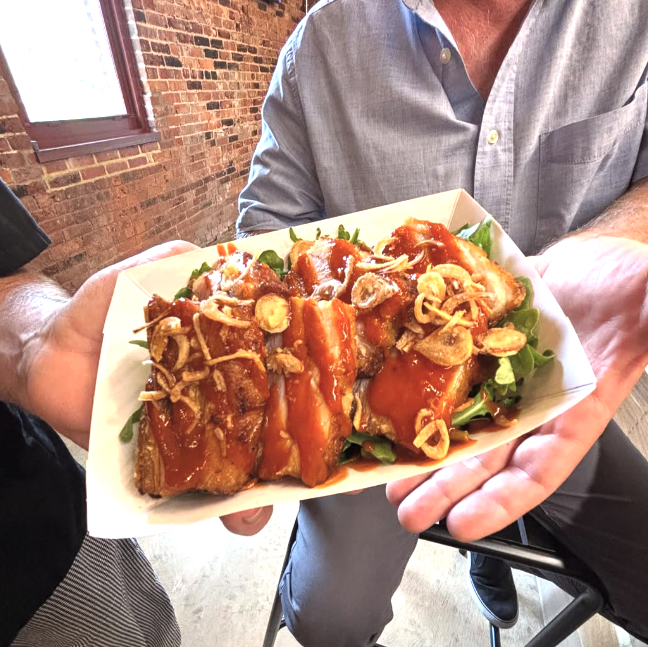 Person holding a tray of barbecue ribs topped with fried onions and sauce, served on a bed of greens.