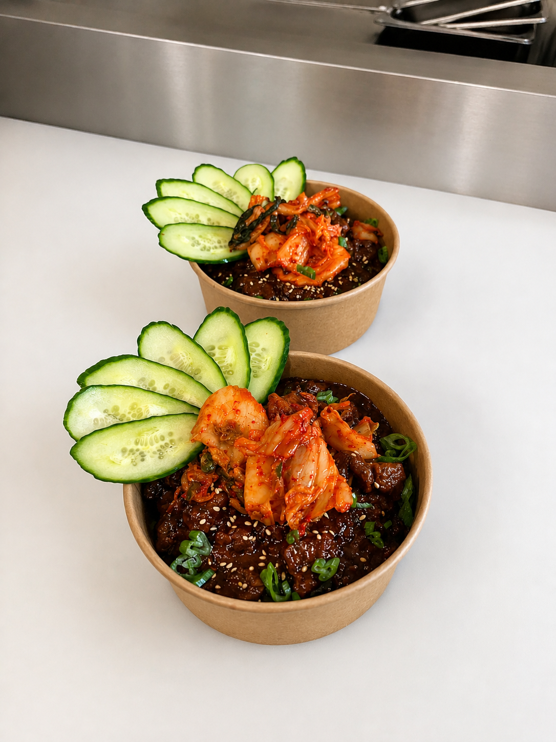 Two brown paper bowls filled with kimchi fried rice, garnished with sliced cucumber, chopped green onions, and sesame seeds, set on a white surface with a metal countertop in the background.