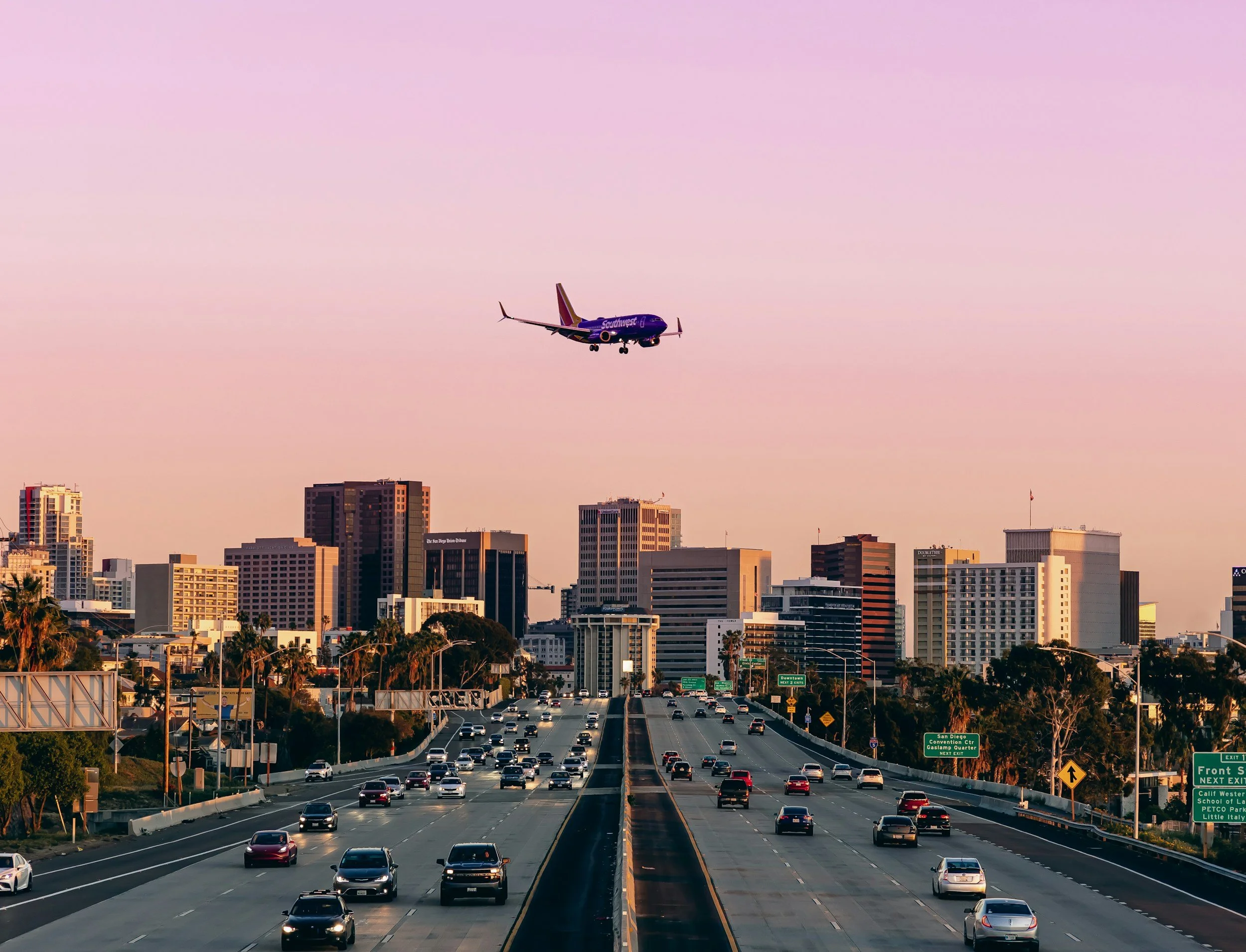 An airplane with Southwest Airlines livery flying over a city skyline during sunset, with high-rise buildings and a busy highway below.