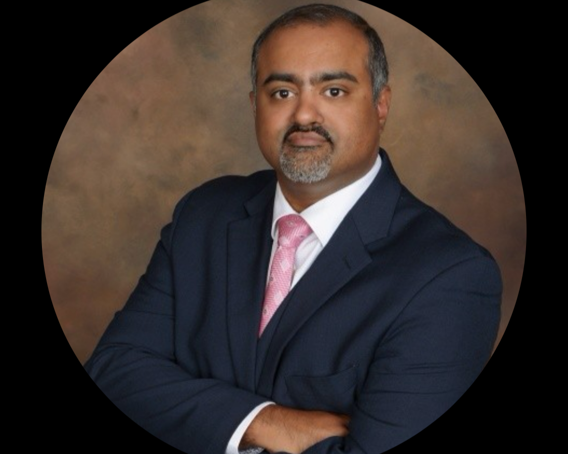 A professional man in a navy suit with a white shirt and pink tie, arms crossed, posing against a brown background.