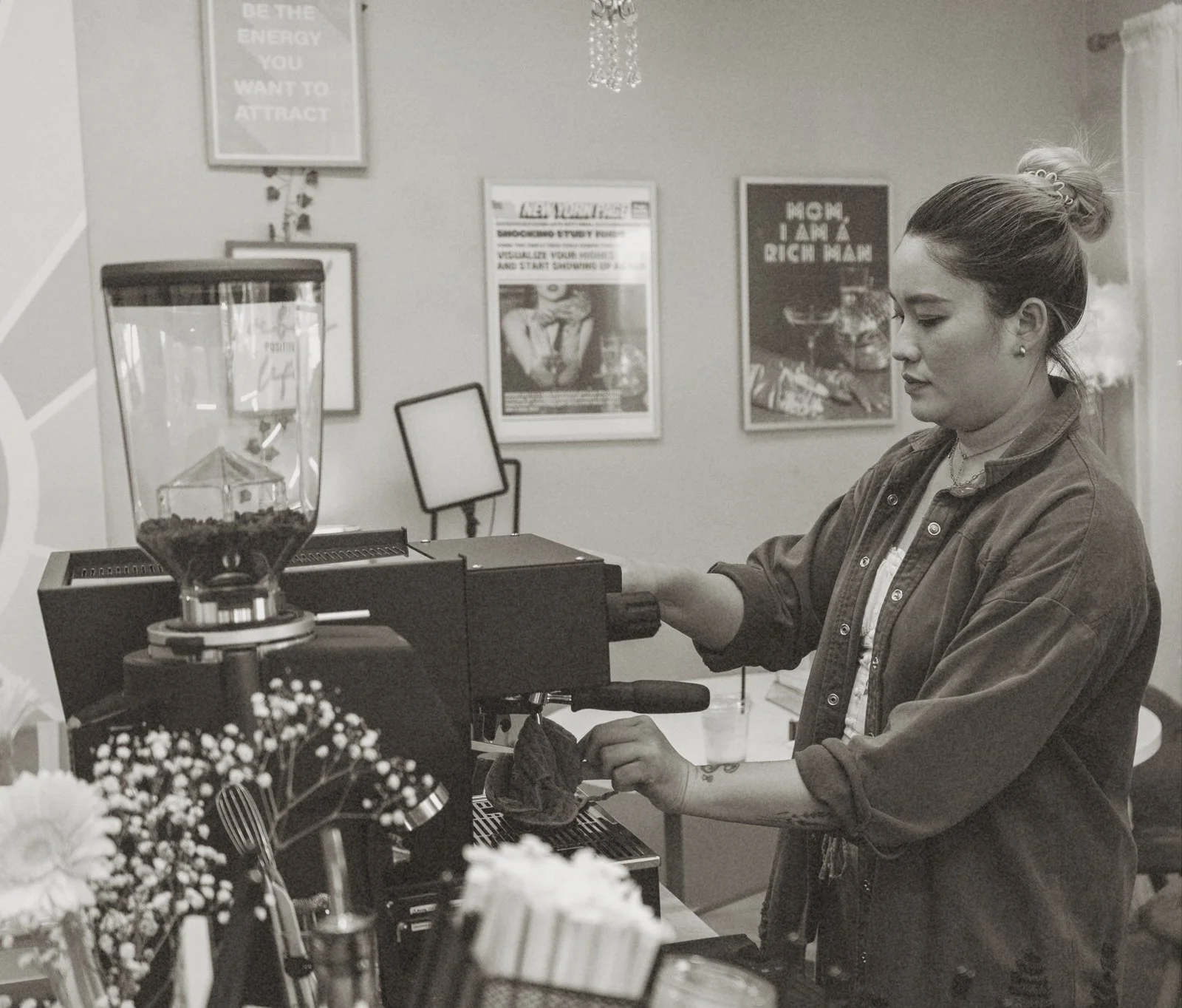 A young woman in a denim jacket and hair in a bun working behind a coffee bar, operating a coffee grinder and espresso machine in a cafe. Behind her are framed posters on the wall, including one that says 'Mom, I am a rich man' and another with a woman posing.