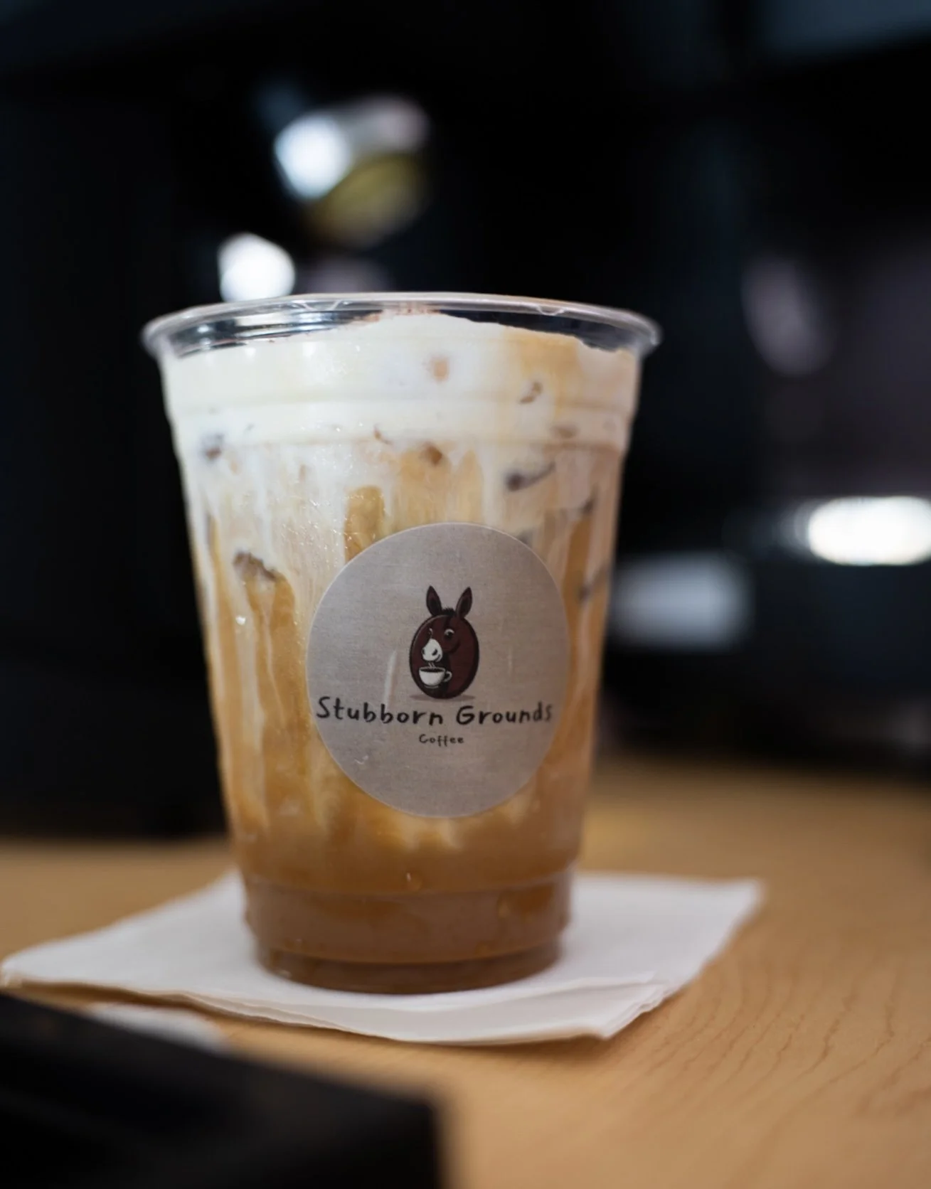 A plastic cup of iced coffee with cream, labeled 'Stubborn Grounds Coffee,' placed on a napkin on a wooden surface.