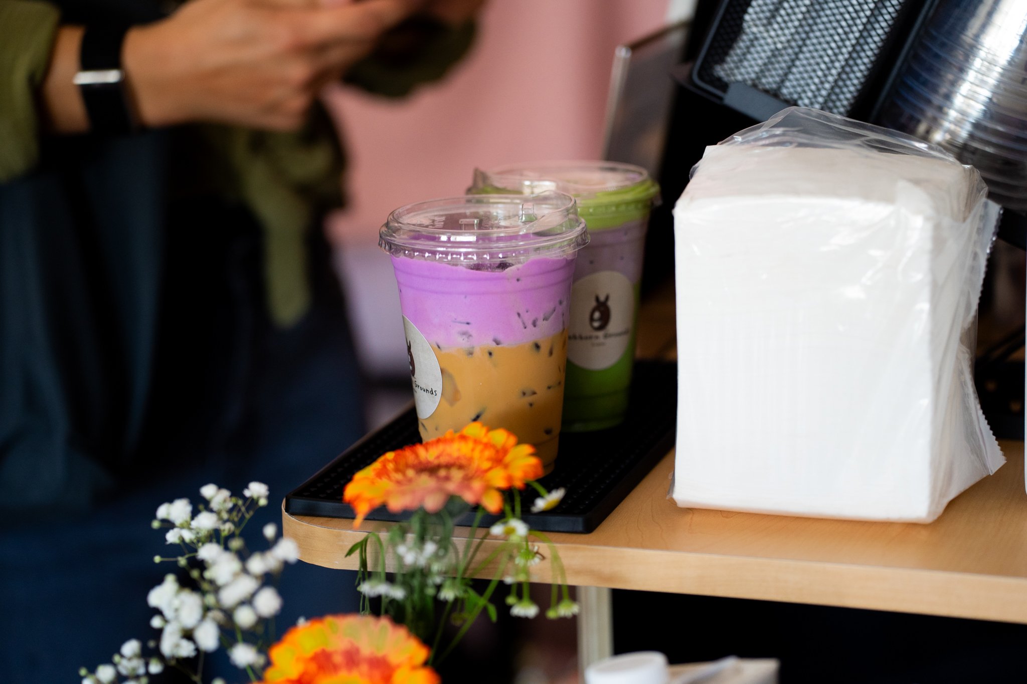 Close-up of a display with colorful bubble tea drinks, a stack of napkins, and some flowers in the foreground.