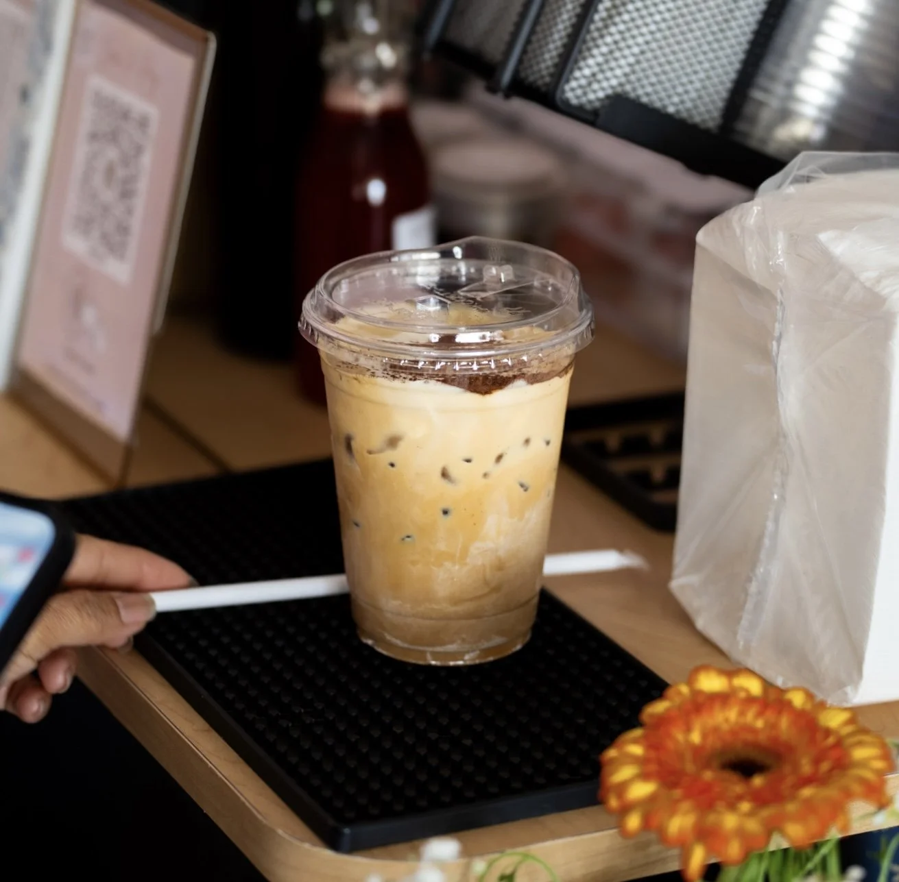Ice coffee drink in a clear plastic cup with a lid, on a black rubber mat at a cafe counter.
