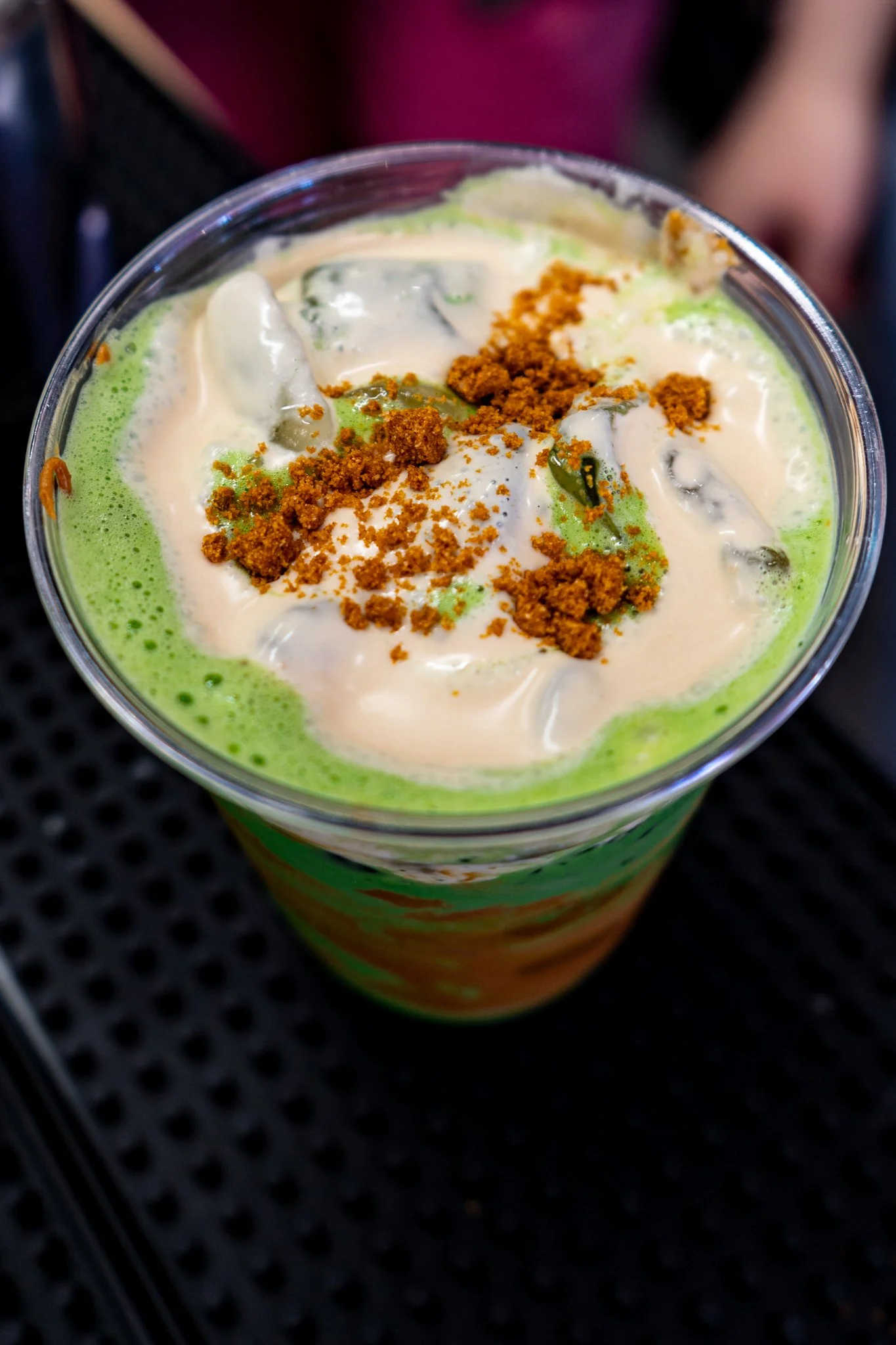 Close-up of a colorful drink with green and white foam, topped with crumbled brown topping and a slice of lime in a clear plastic cup.