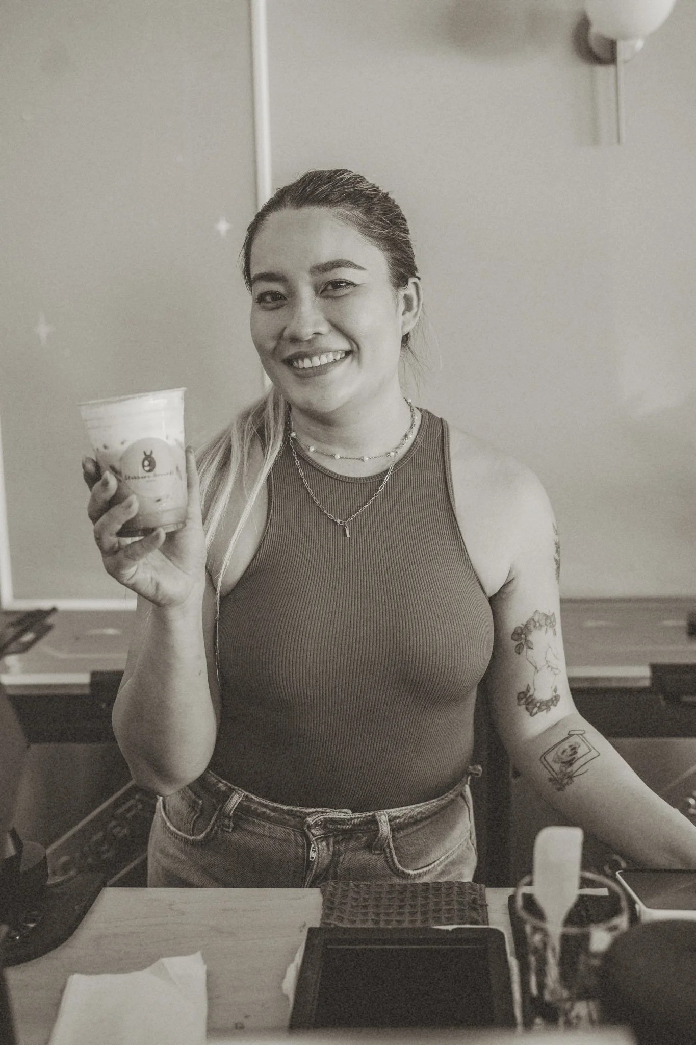 A woman smiling in a mirror, holding a cup with a logo on it, wearing a sleeveless top and jeans, showcasing tattoos on her arm.