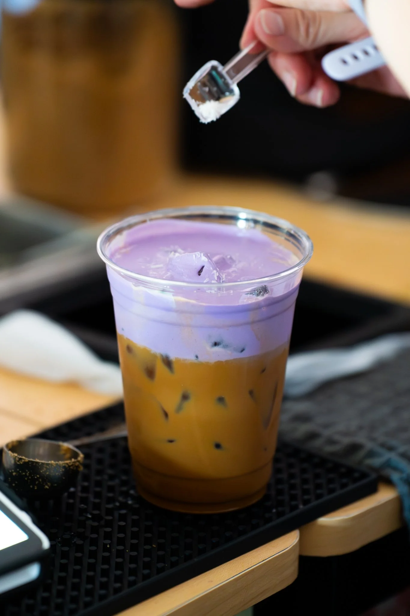 Close-up of a plastic cup filled with purple and yellow layered drink with ice, on a black tray, with a person using a dropper or pipette to add a white substance on top.