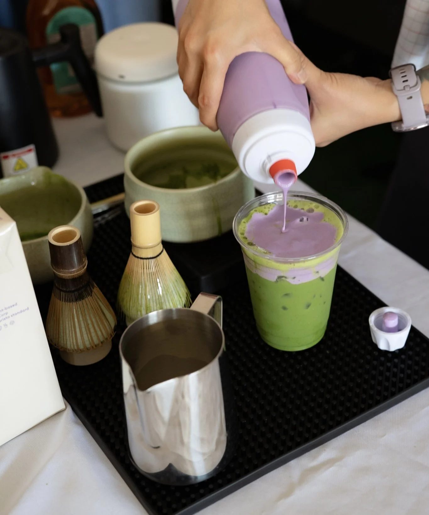 A person pouring purple liquid into a green tea latte with ice, on a black tray with small ceramic cups, a metal creamer pitcher, and tea brewing utensils, with a white tablecloth in the background.