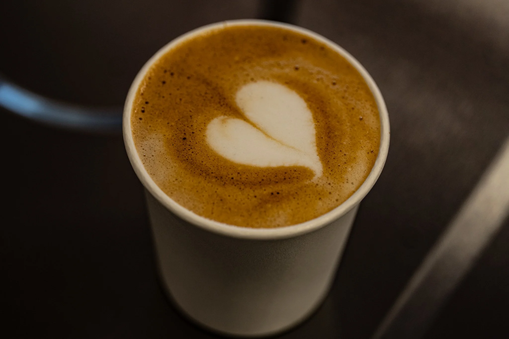 Top view of a paper cup filled with coffee that has a heart-shaped latte art design on the foam, on a dark surface.