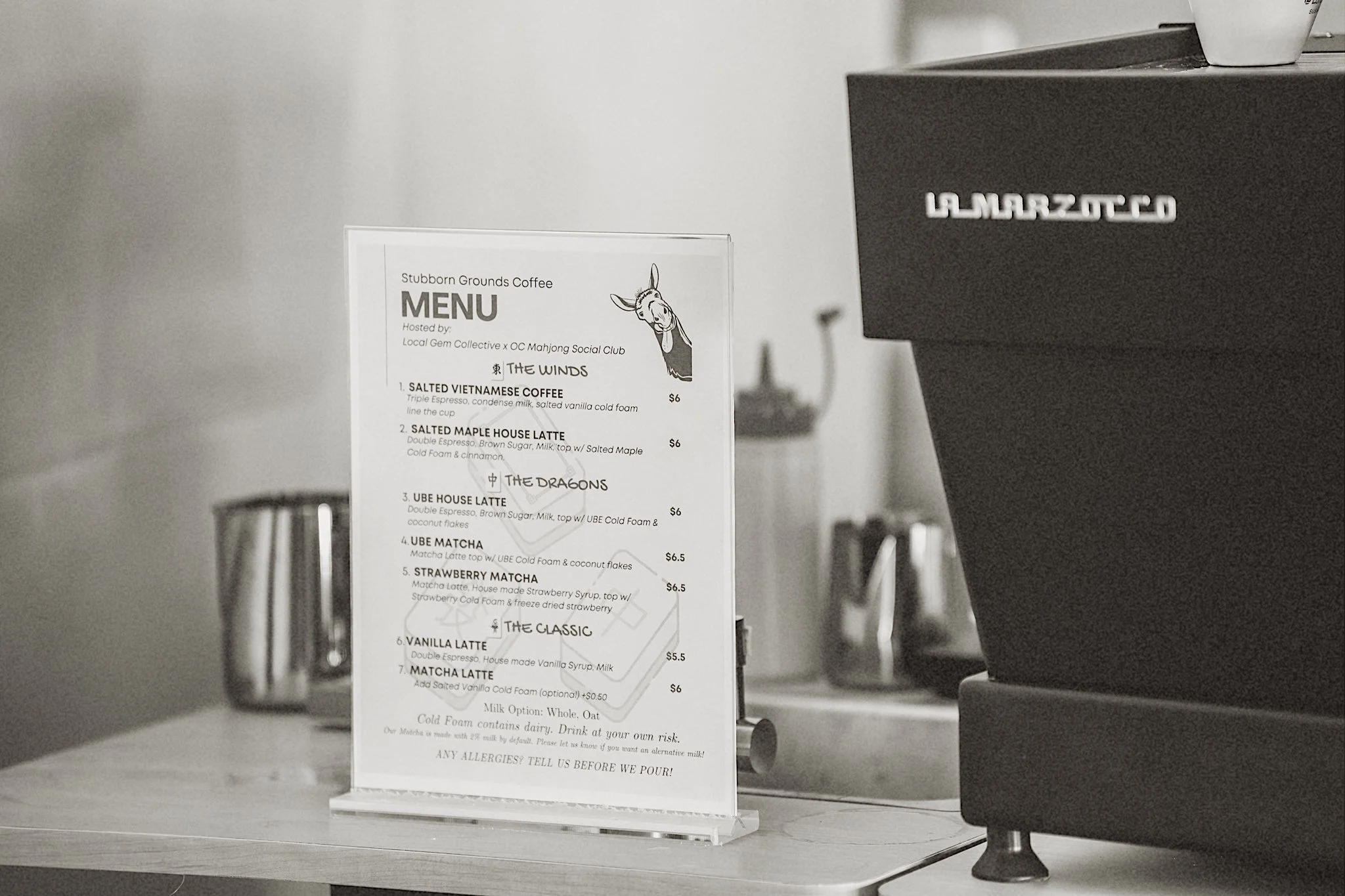 A menu from Stubborn Grounds Coffee positioned on a clear stand on a table, with a coffee machine to the right. The menu features various coffee drinks and prices, along with a cartoon donkey illustration and a list of coffee options.
