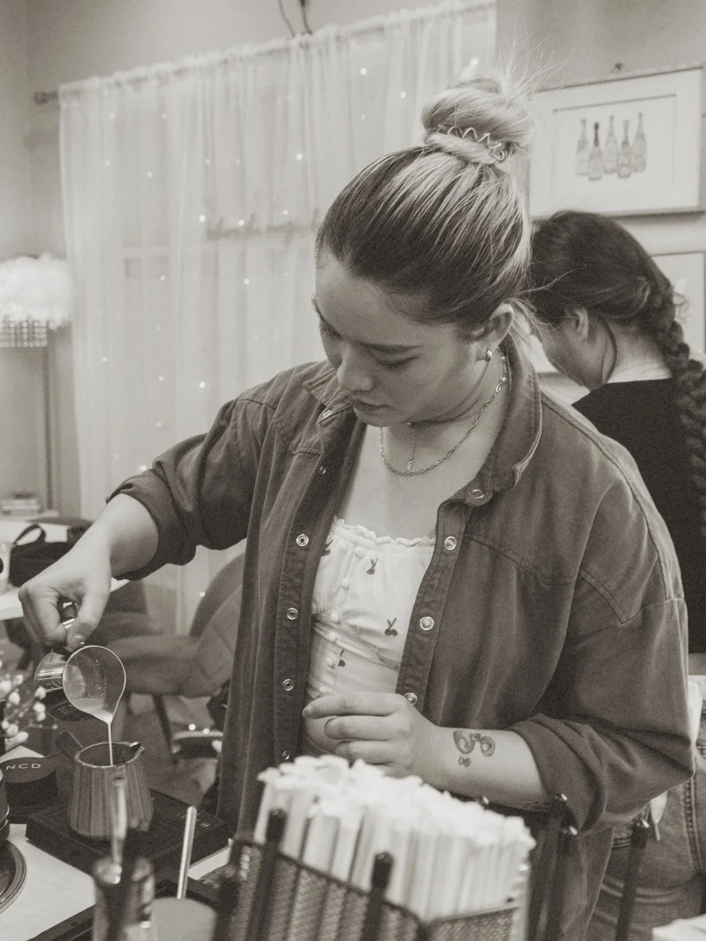 A woman with blonde hair tied up in a bun, wearing a denim shirt, streams liquid into a cocktail shaker at a bar or coffee station. She has a tattoo on her left forearm and is focused on her task. Another woman with dark hair in a braid is in the background.