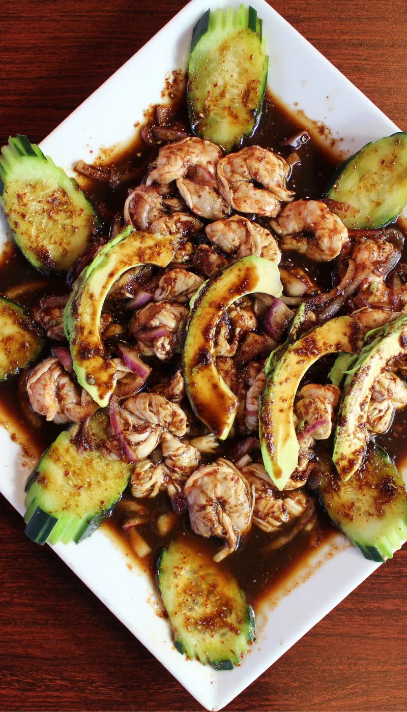 A rectangular white plate with cooked shrimp, cucumber slices, and sauce, garnished with chopped red onions.